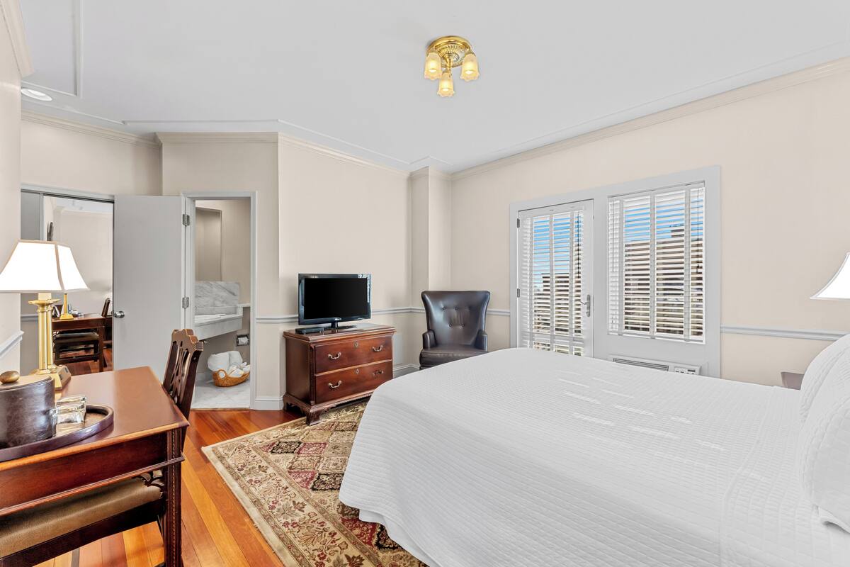 A hotel room with a large bed, a desk with a lamp, a television, a chair, and a wardrobe.