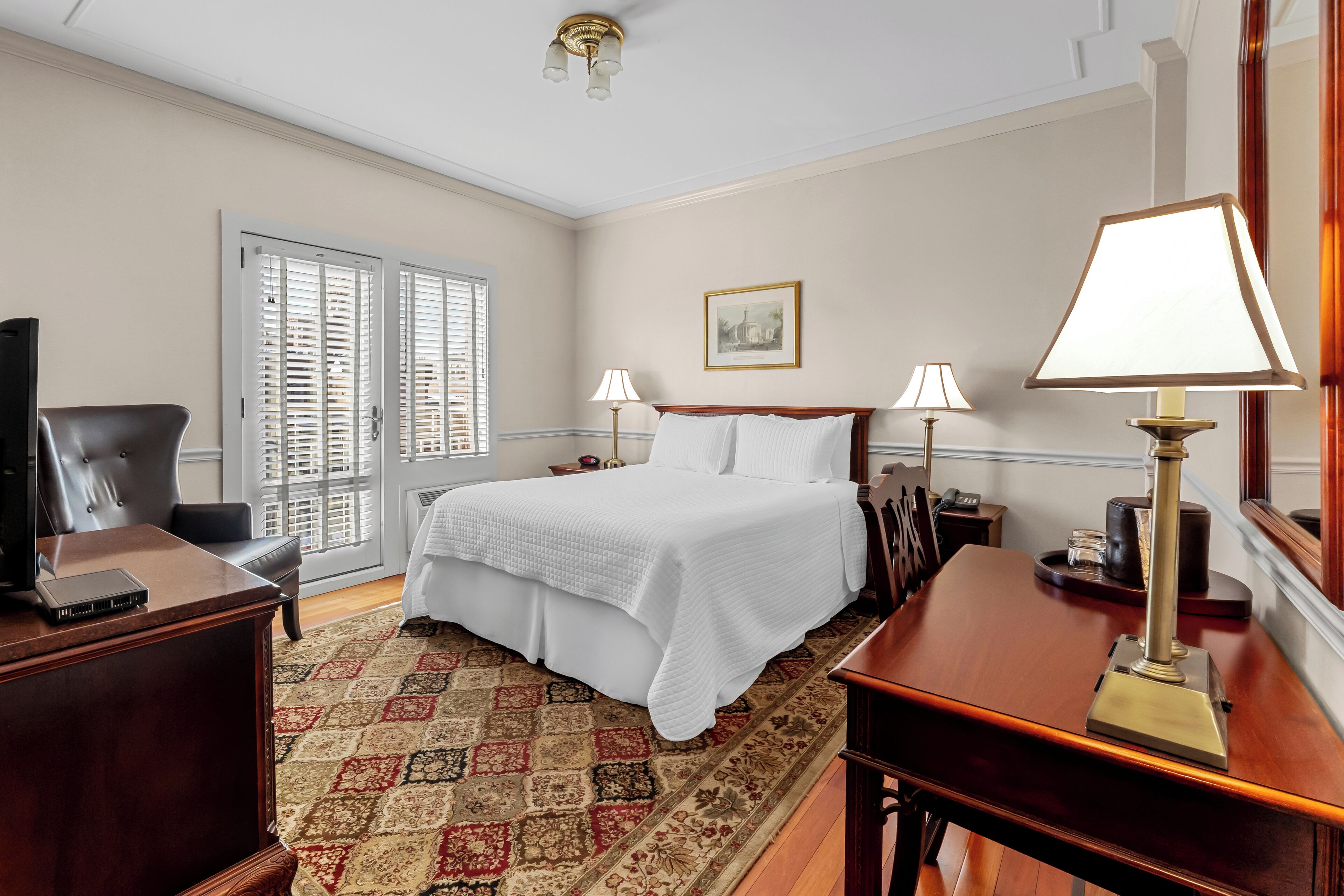 A hotel room with a large bed, two bedside lamps, a desk with a lamp, a chair, a television, and a patterned rug.