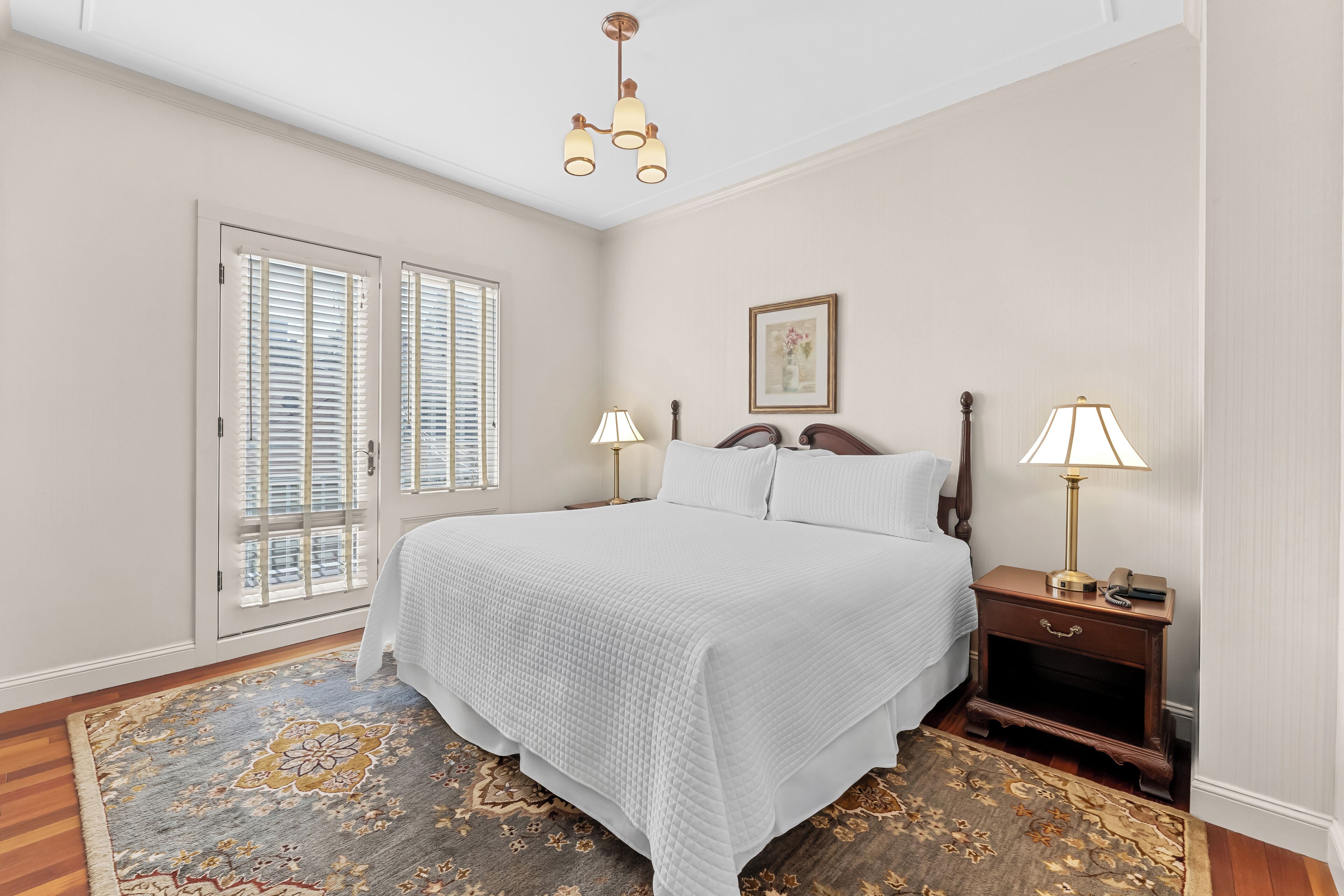 A bedroom with a large bed, two bedside lamps, a nightstand, a framed picture on the wall, and a patterned rug.