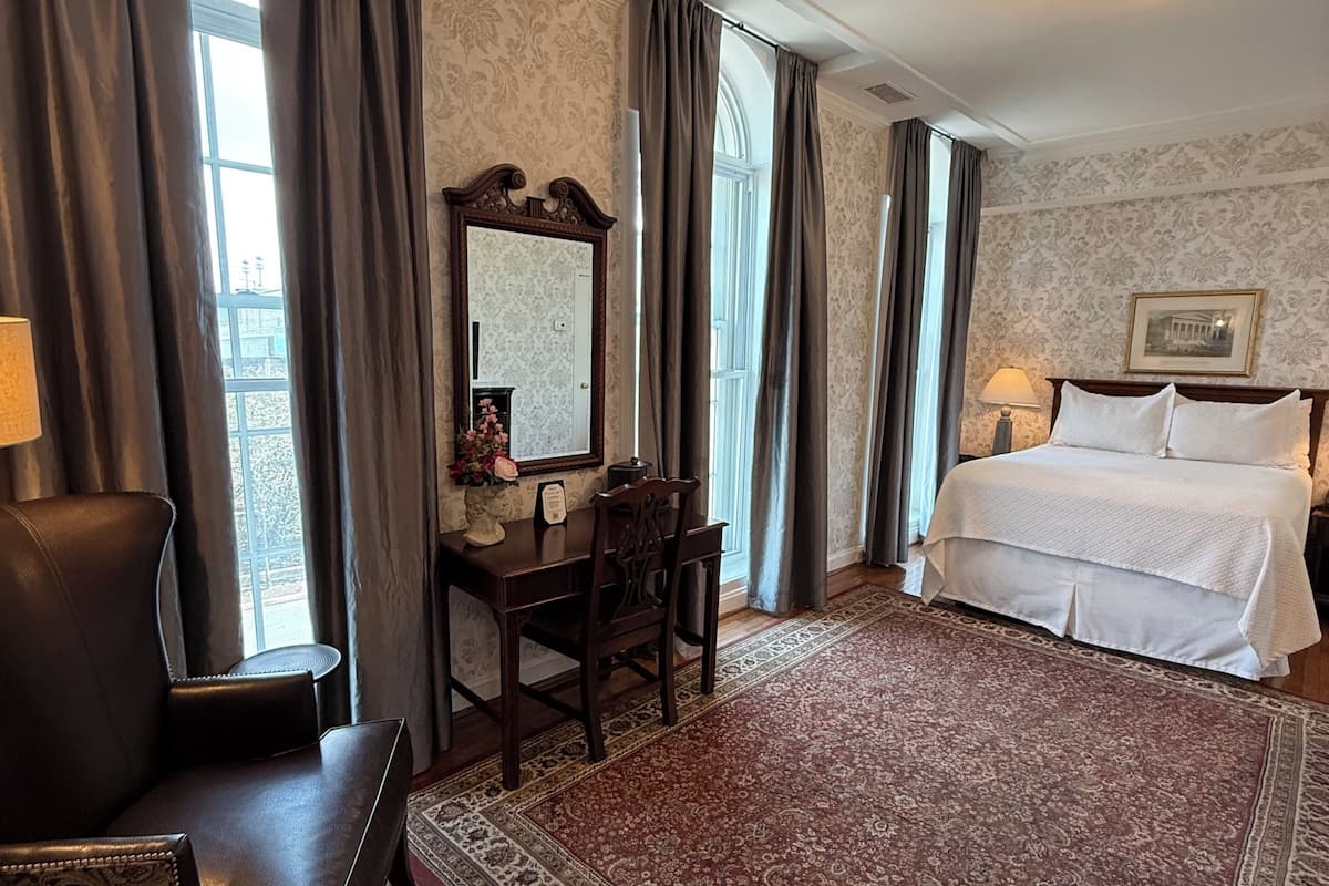 A hotel room with a bed, a desk with a chair, a large mirror, and a patterned rug.