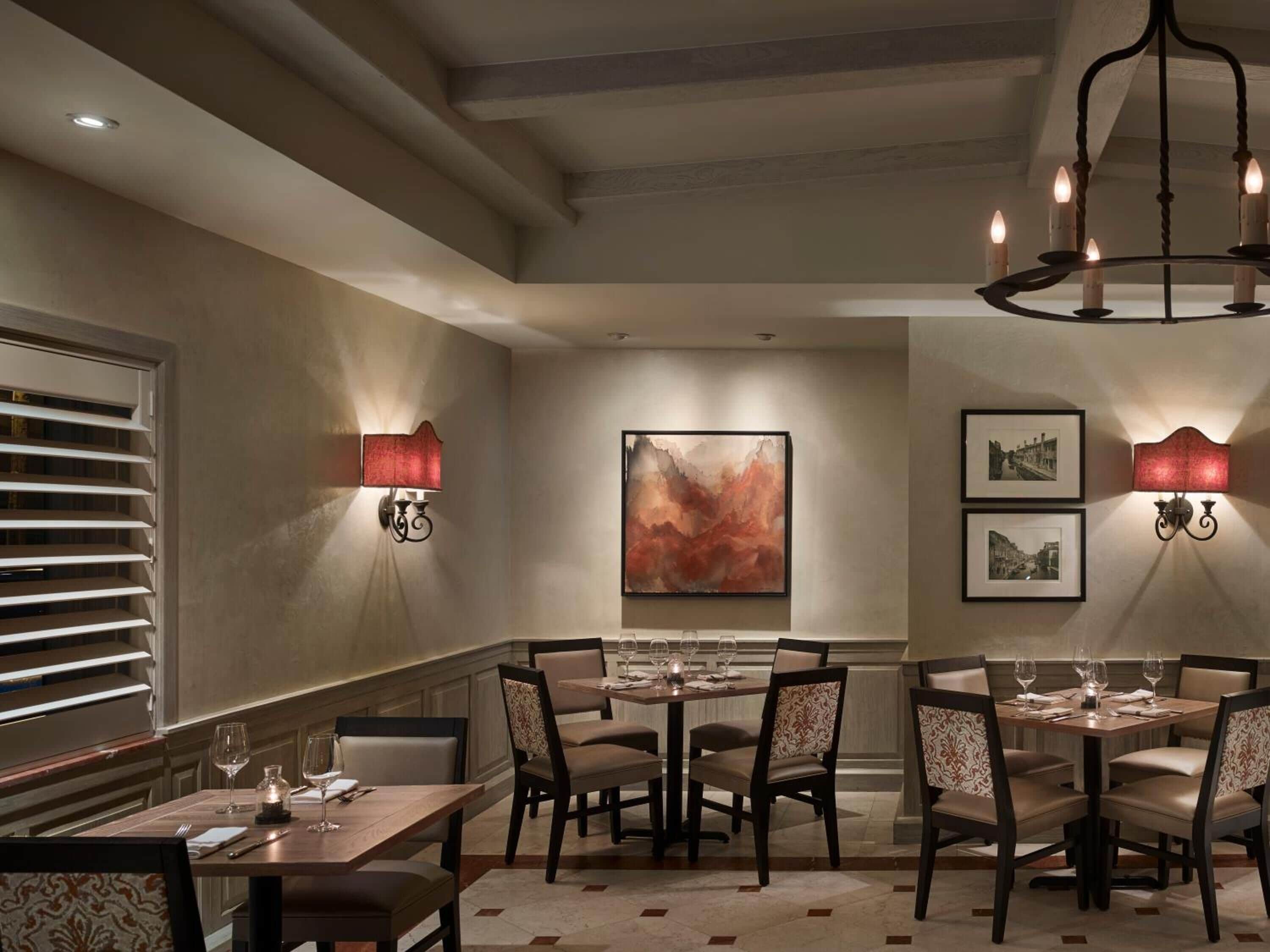 A dining area with tables set for four, wall-mounted sconces, and a large abstract painting on the wall.