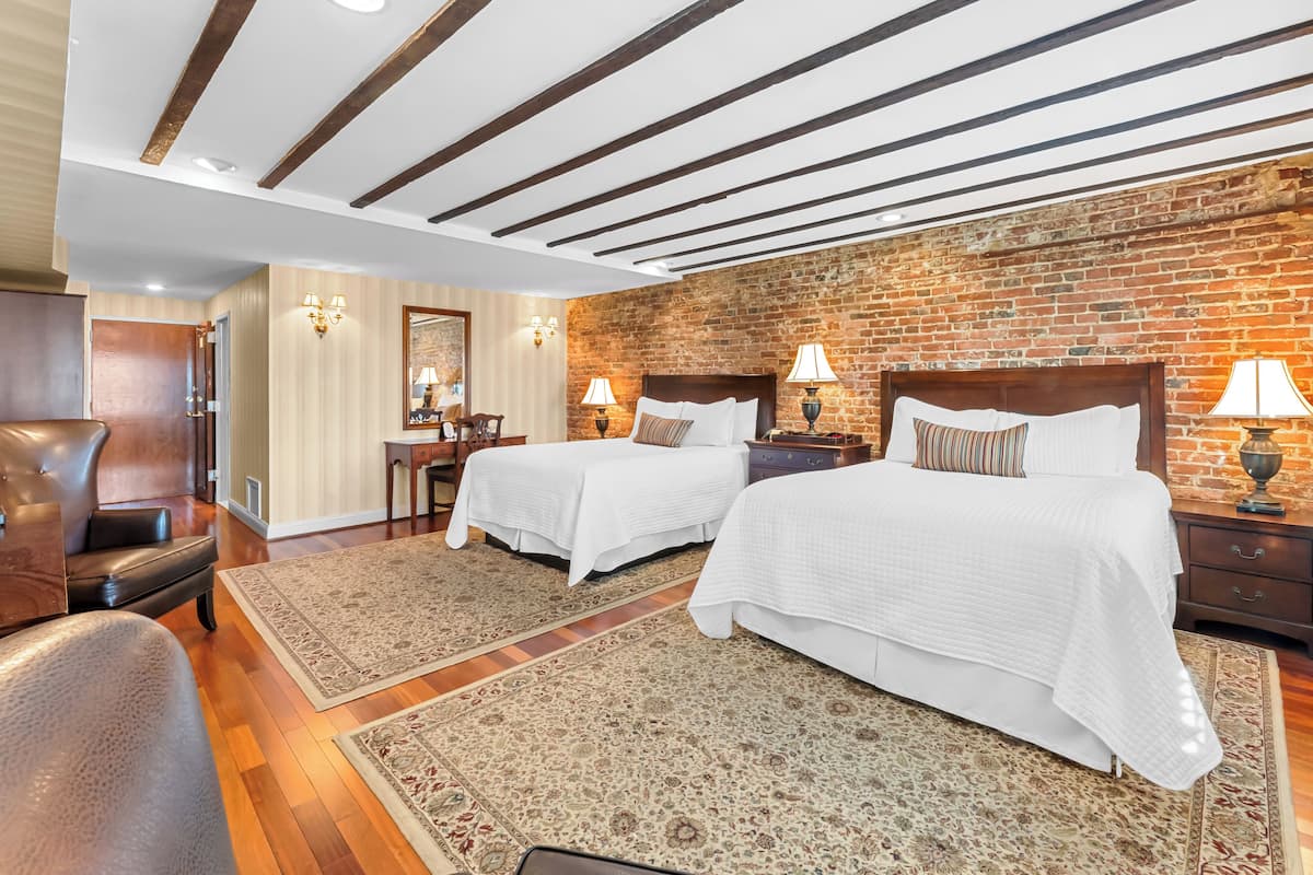 A hotel room with two beds, a brick wall, wooden beams on the ceiling, and a large rug.