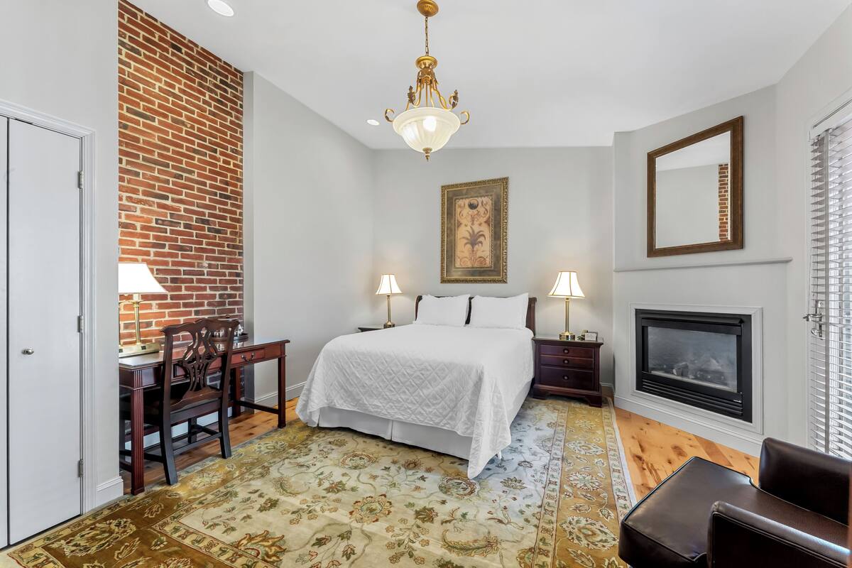 A bedroom with a brick accent wall, a chandelier, a fireplace, and a bed with a white quilt.