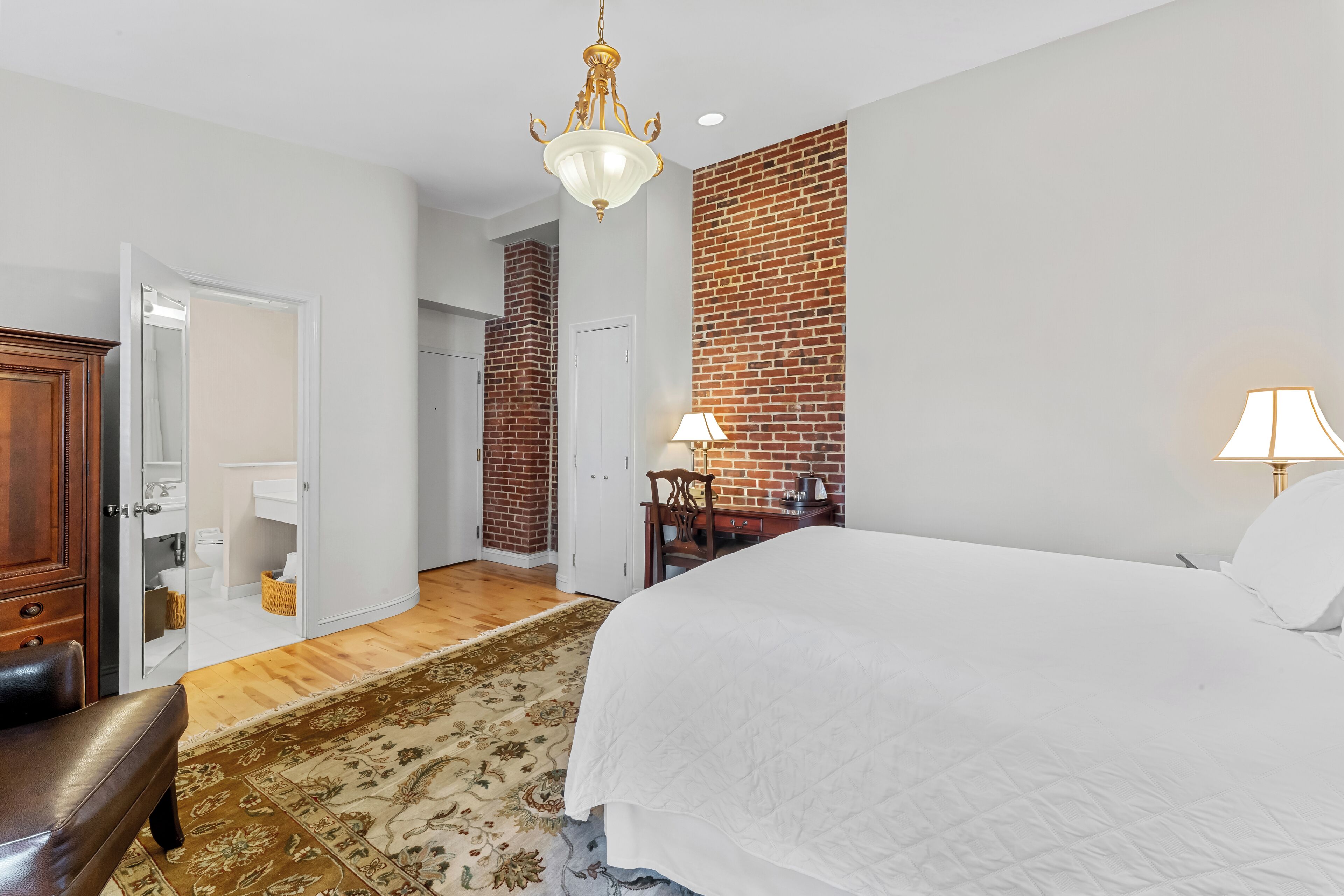 A bedroom with a large bed, a wooden bench, a desk with a lamp, and a brick accent wall.