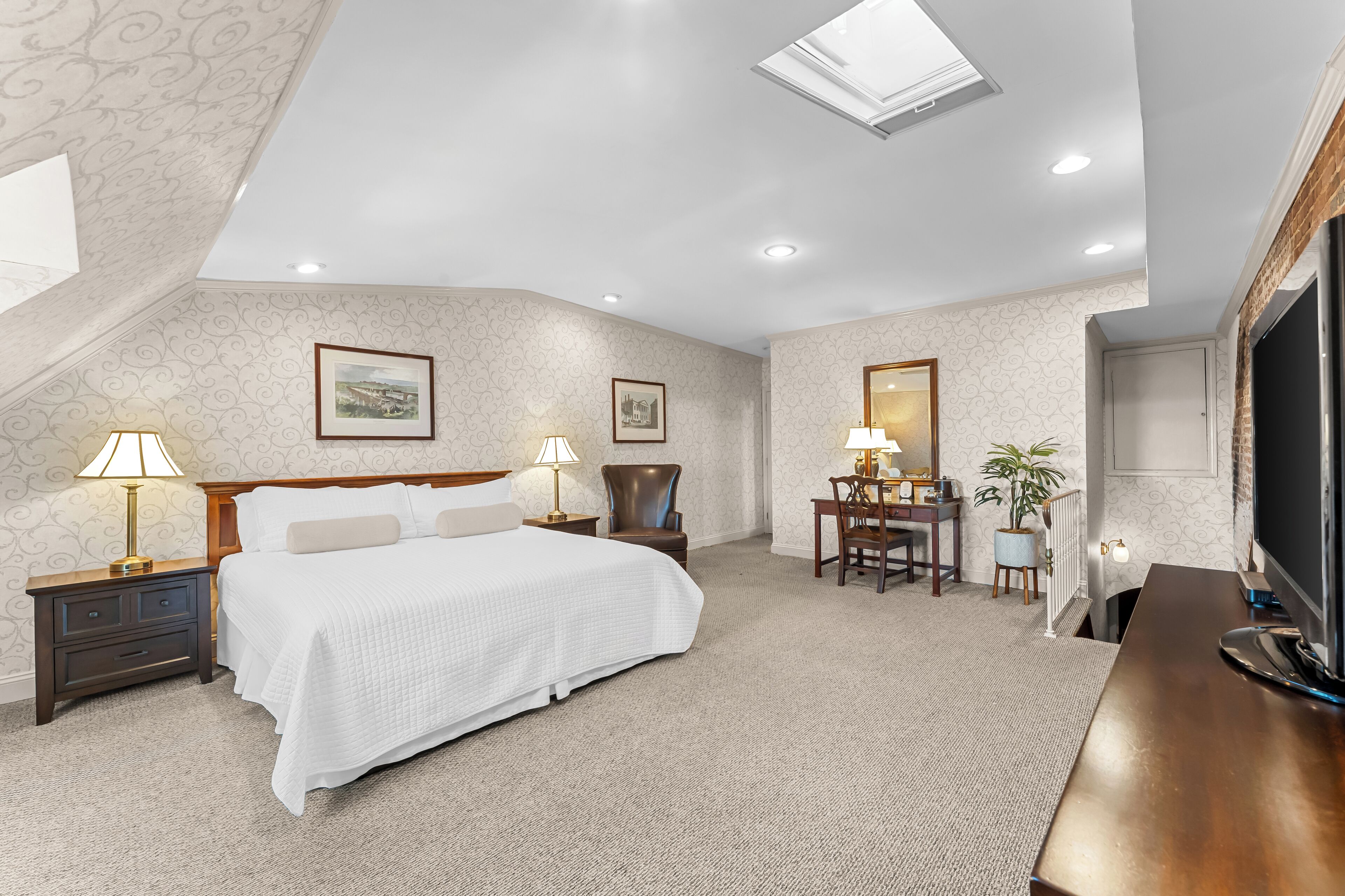 A bedroom with a large bed, two bedside tables with lamps, a desk with a chair, a television, and a skylight.