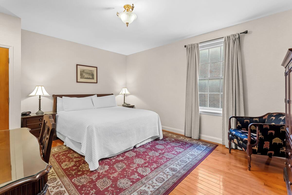 A bedroom with a large bed, two bedside lamps, a wooden dresser, a desk, and a patterned rug.