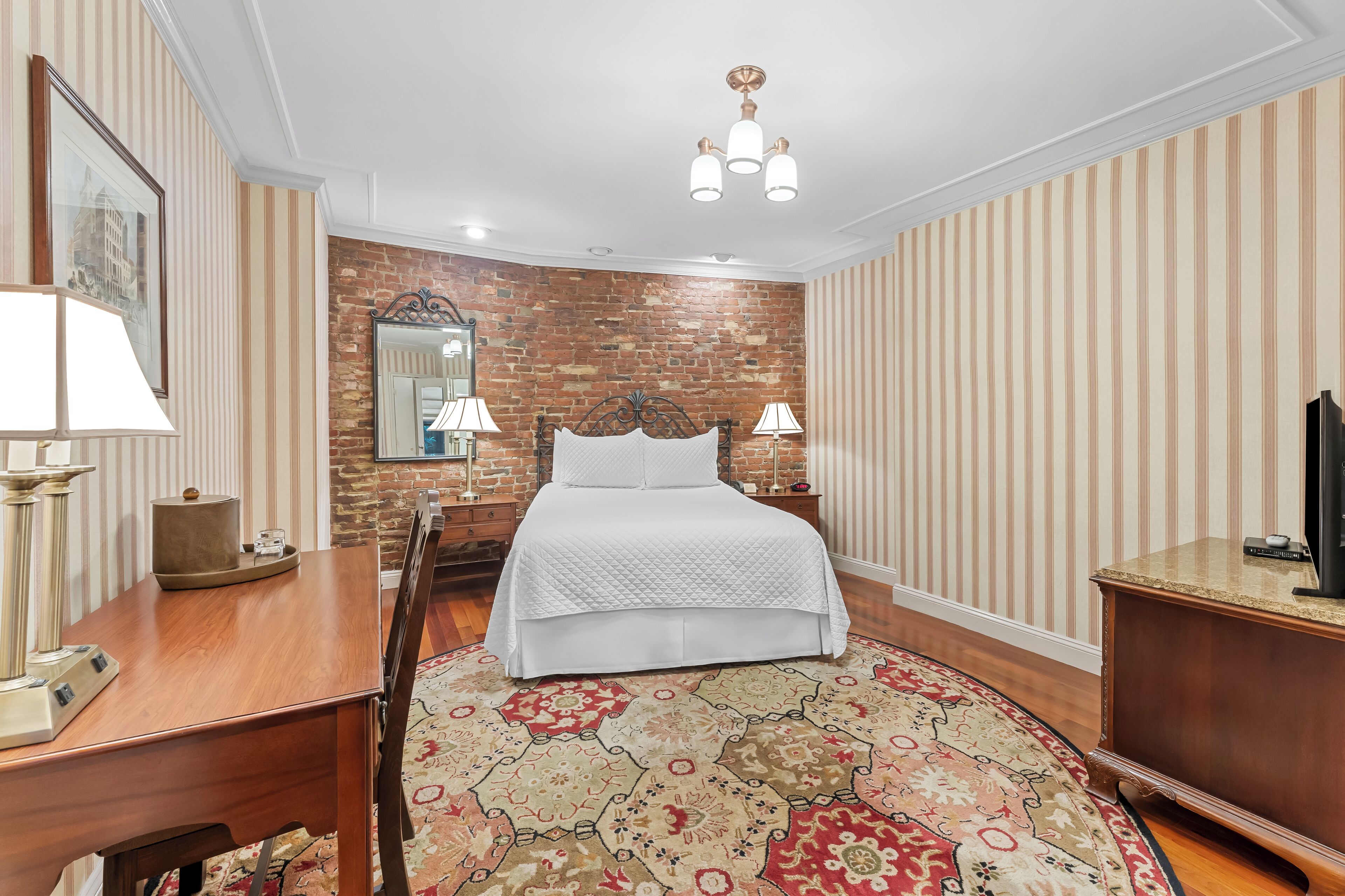 A bedroom with a brick wall, a bed with white bedding, two bedside lamps, a wooden dresser, a mirror, and a patterned rug.