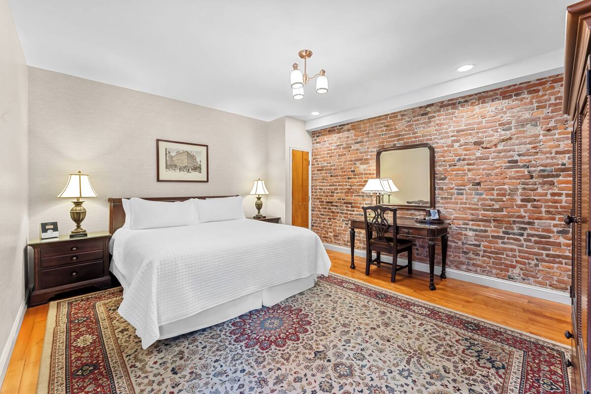 A bedroom with a brick wall, a bed with white bedding, two bedside tables with lamps, a wooden desk with a chair, and a patterned rug.