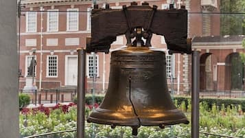 A large bronze bell with inscriptions, mounted on a structure with a broken top, set against a backdrop of a brick building with windows and a statue.