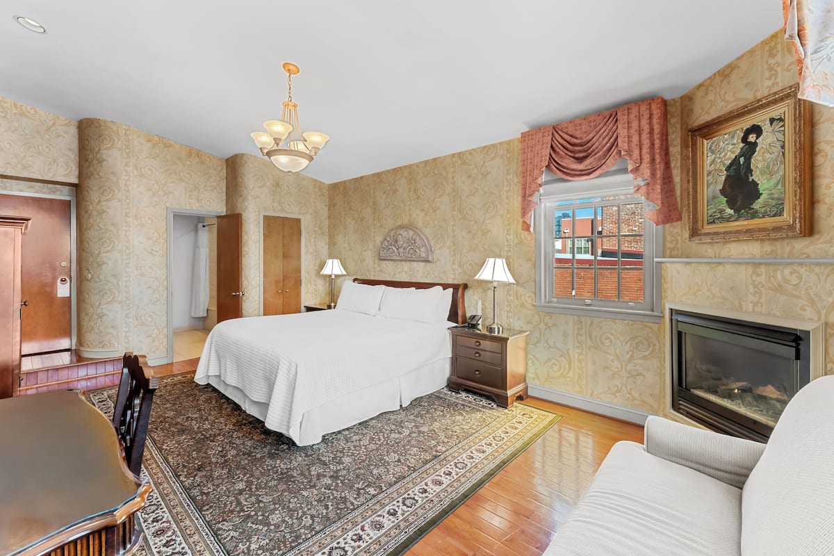 A bedroom with a large bed, two bedside tables with lamps, a fireplace, a dining table, and a patterned rug.