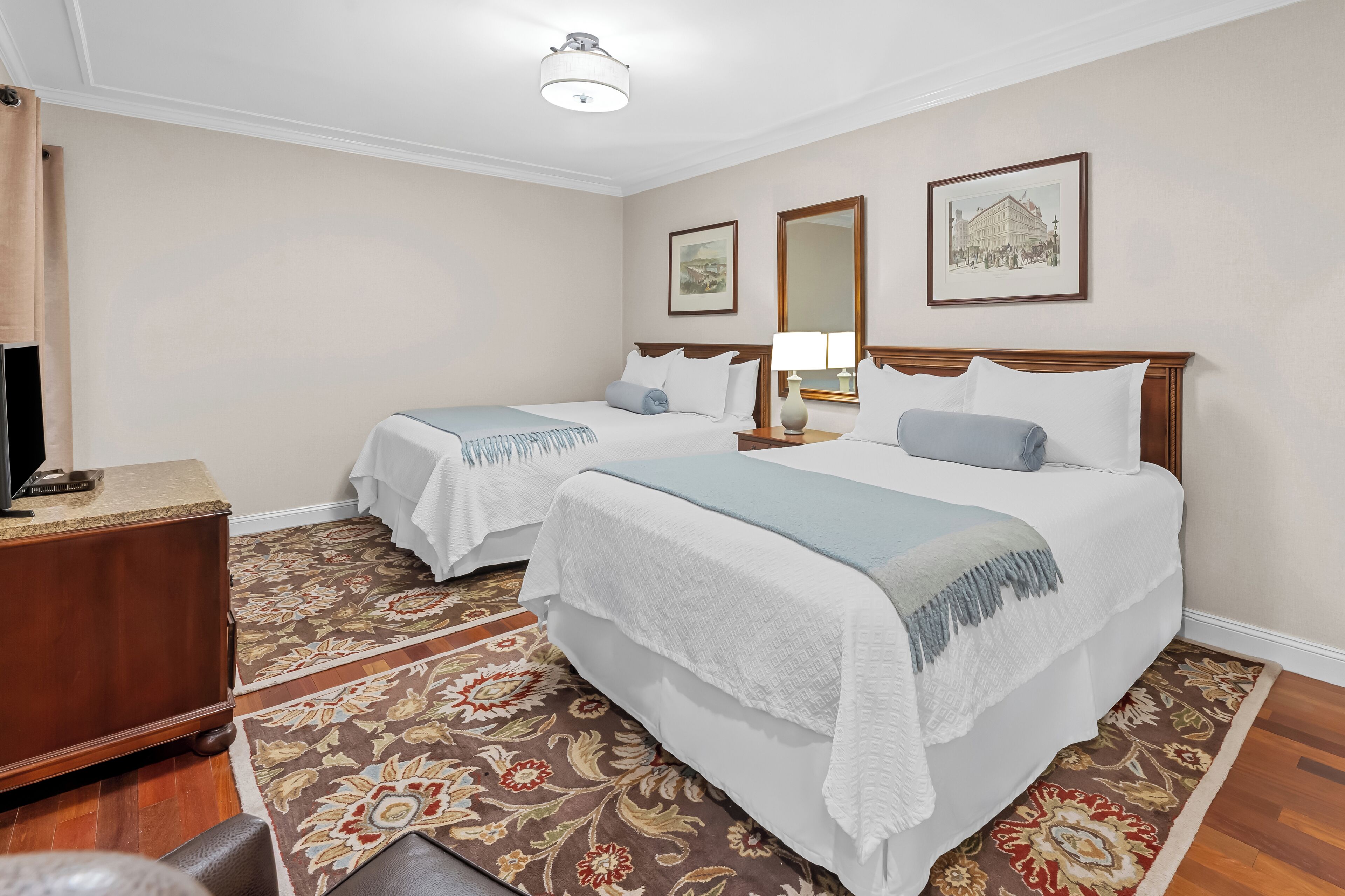 A hotel room with two beds, a wooden nightstand, a desk with a lamp, and a patterned rug.