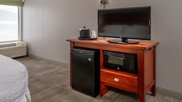 A hotel room with a bed, a small kitchenette with a refrigerator, microwave, and coffee maker, and a flat-screen TV on a wooden stand.
