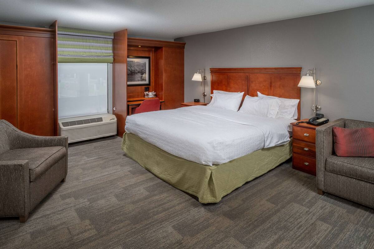 A hotel room with a large bed, two bedside tables, a desk with a chair, and a window with blinds.