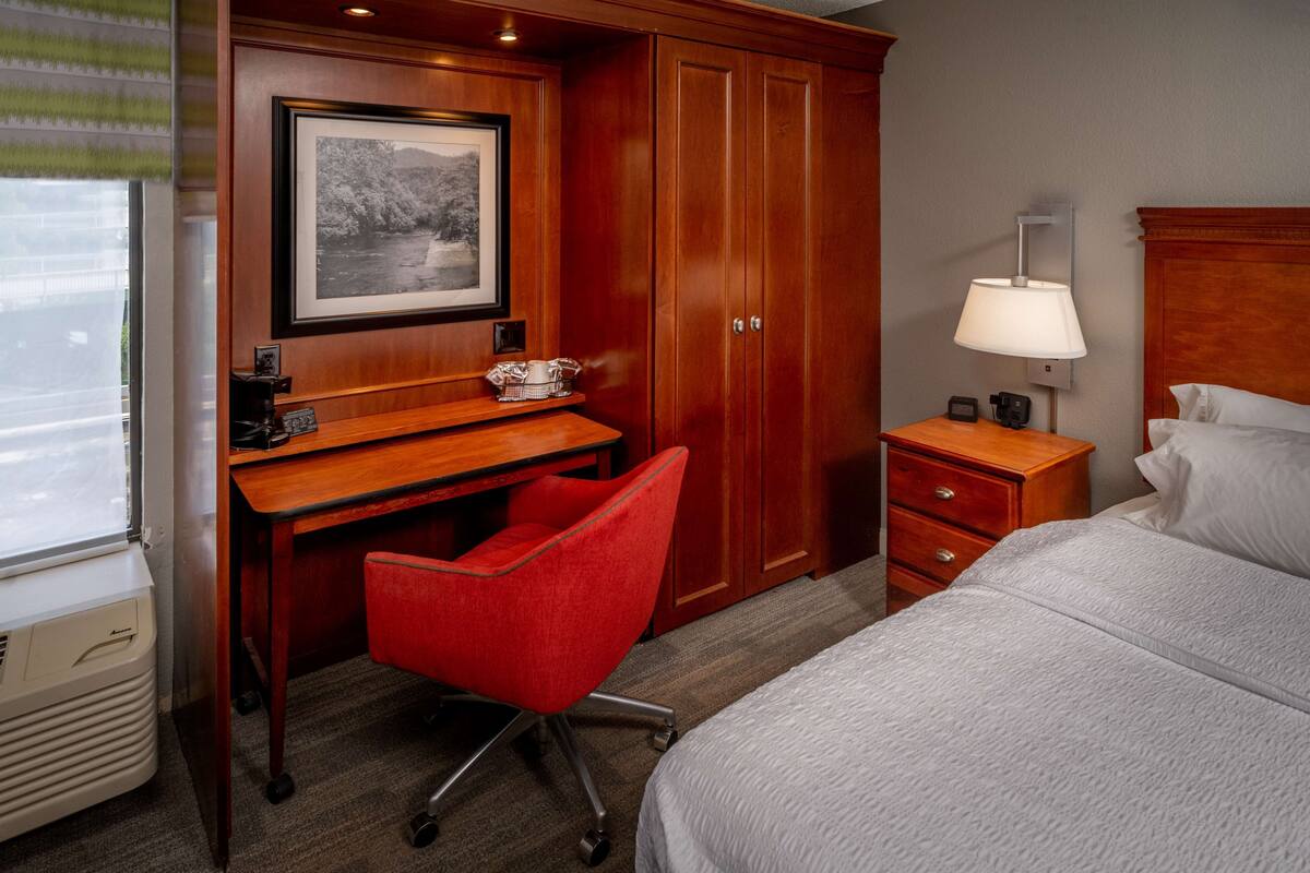 A hotel room with a wooden desk, a red chair, a bed, a nightstand, a lamp, and a framed picture on the wall.