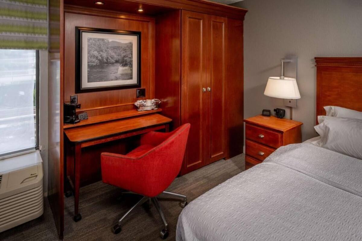Quarto de hotel com escrivaninha de madeira, uma cadeira vermelha, uma cama, mesa de cabeceira e uma luminária.