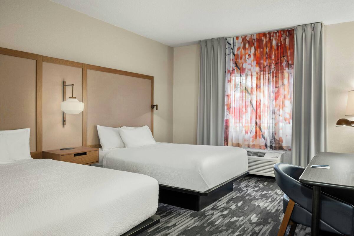 A hotel room with two beds, a desk, and a chair. There is a window with curtains, a wall-mounted lamp, and an air conditioning unit.