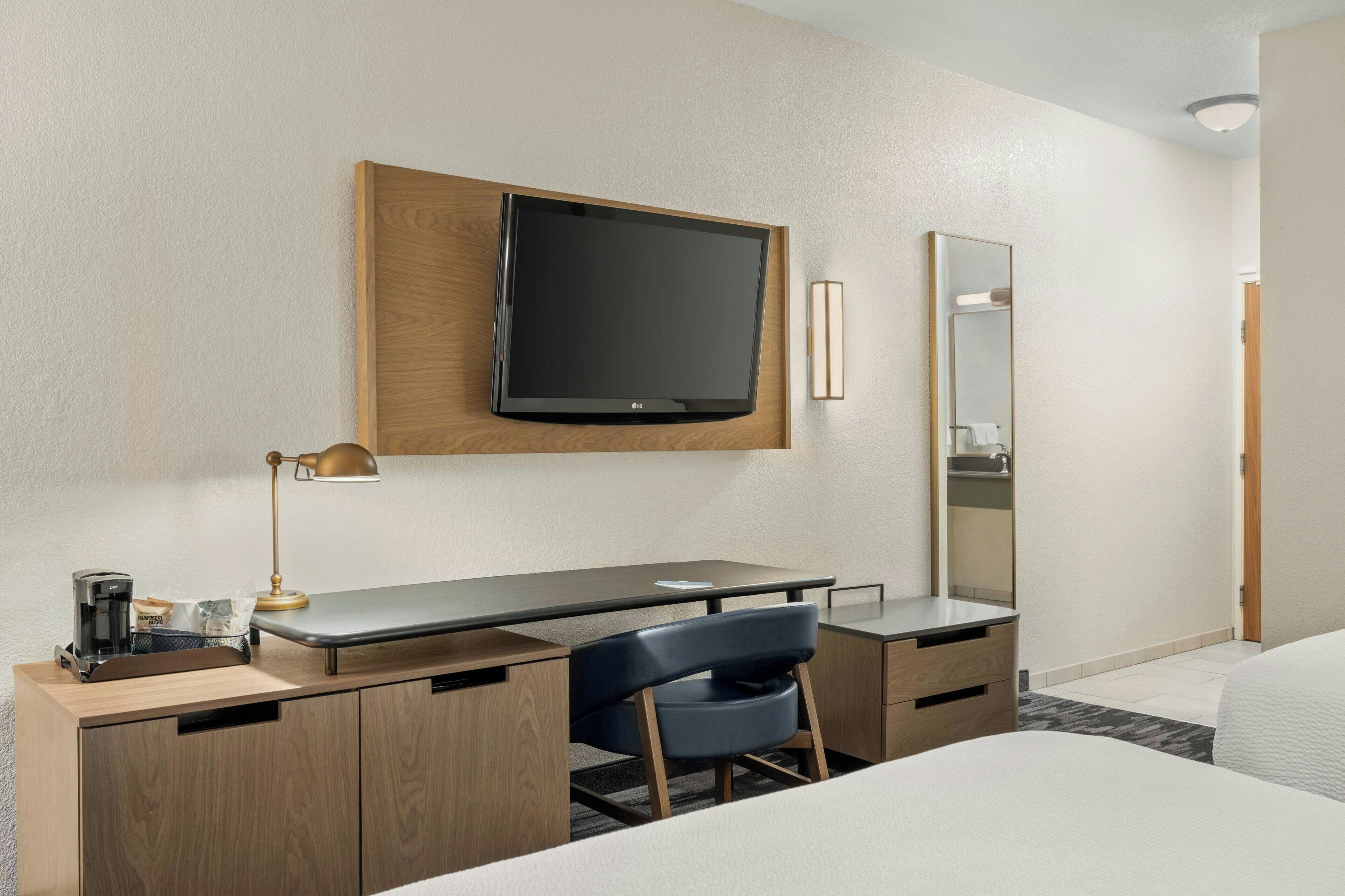 A hotel room with a wooden desk, a flat-screen TV, a mirror, a bed, and a chair.