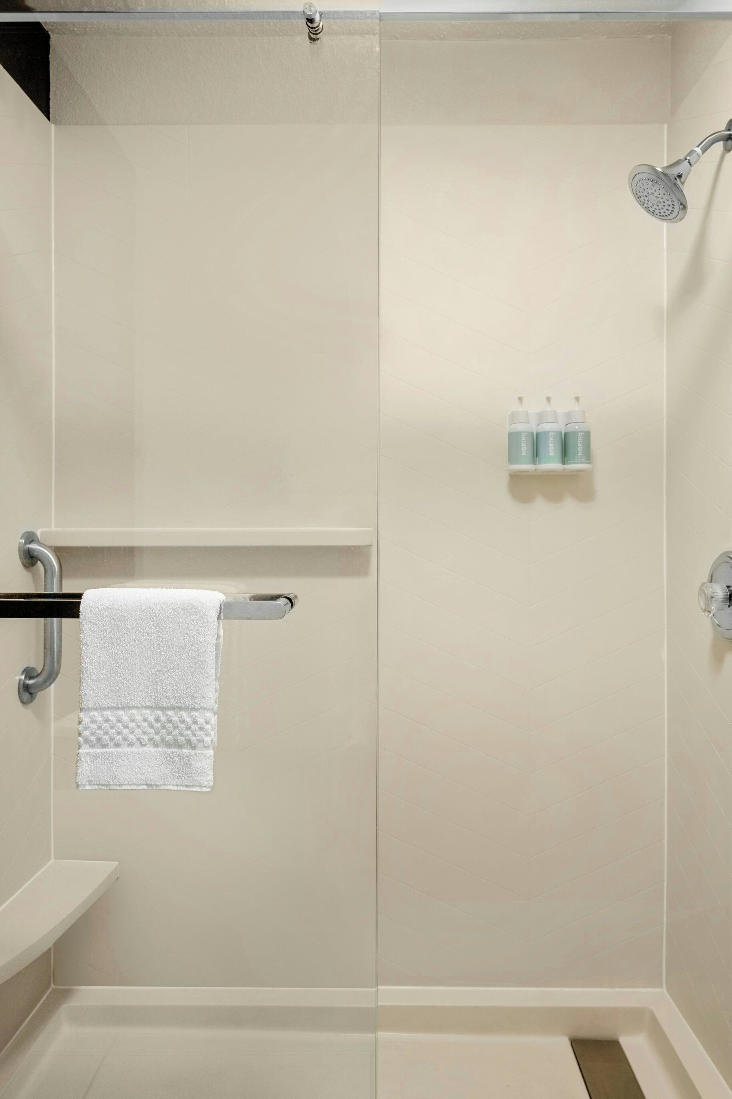 A walk-in shower with a white towel hanging on a metal rack, a handheld showerhead, and a shelf with three bottles of shampoo.