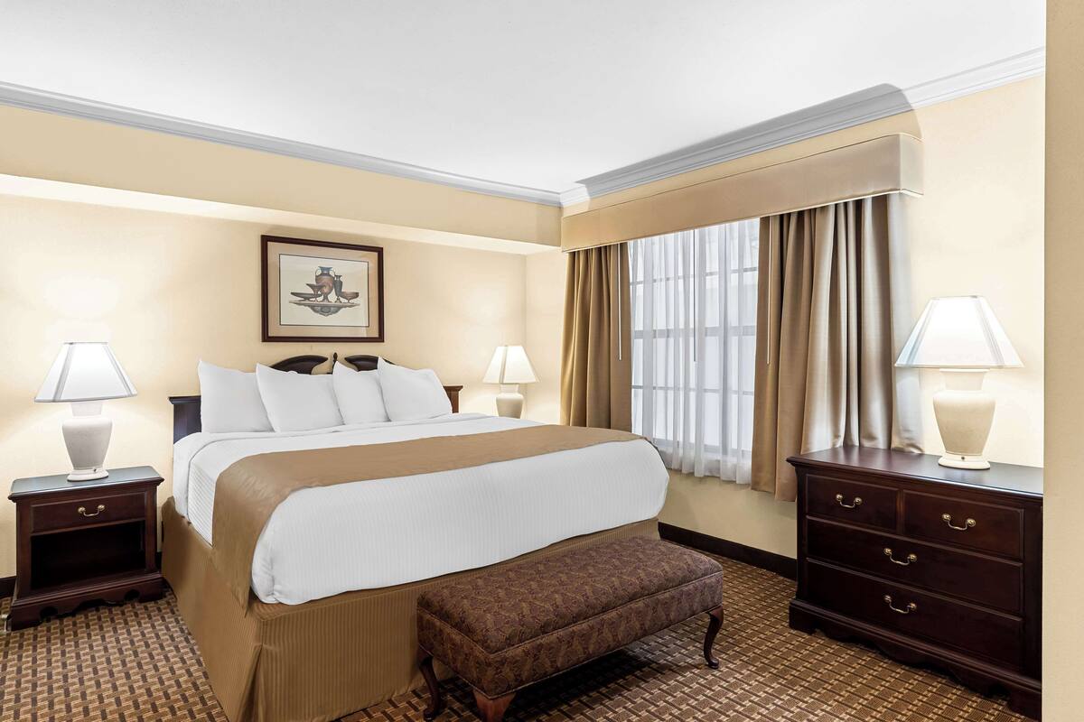 Une chambre d’hôtel avec un grand lit, deux tables de chevet avec des lampes, un banc, un tableau encadré au mur et une fenêtre avec des rideaux.