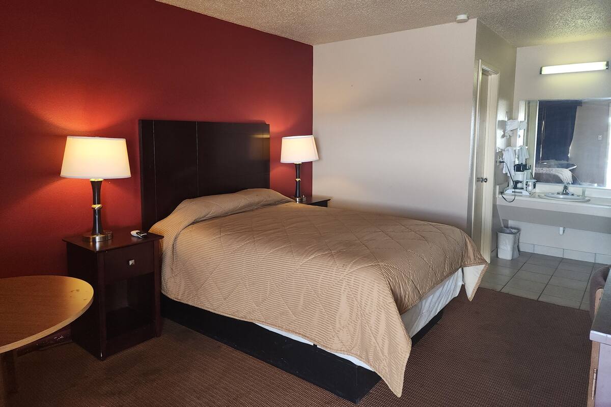 A hotel room with a bed, two bedside lamps, a small table, a bathroom with a sink and mirror, and a red accent wall.