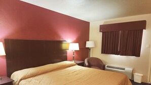 A hotel room with a large bed, a nightstand, a chair, a lamp, a window with curtains, and an air conditioning unit. - Colonial Inn Monahans (Monahans)