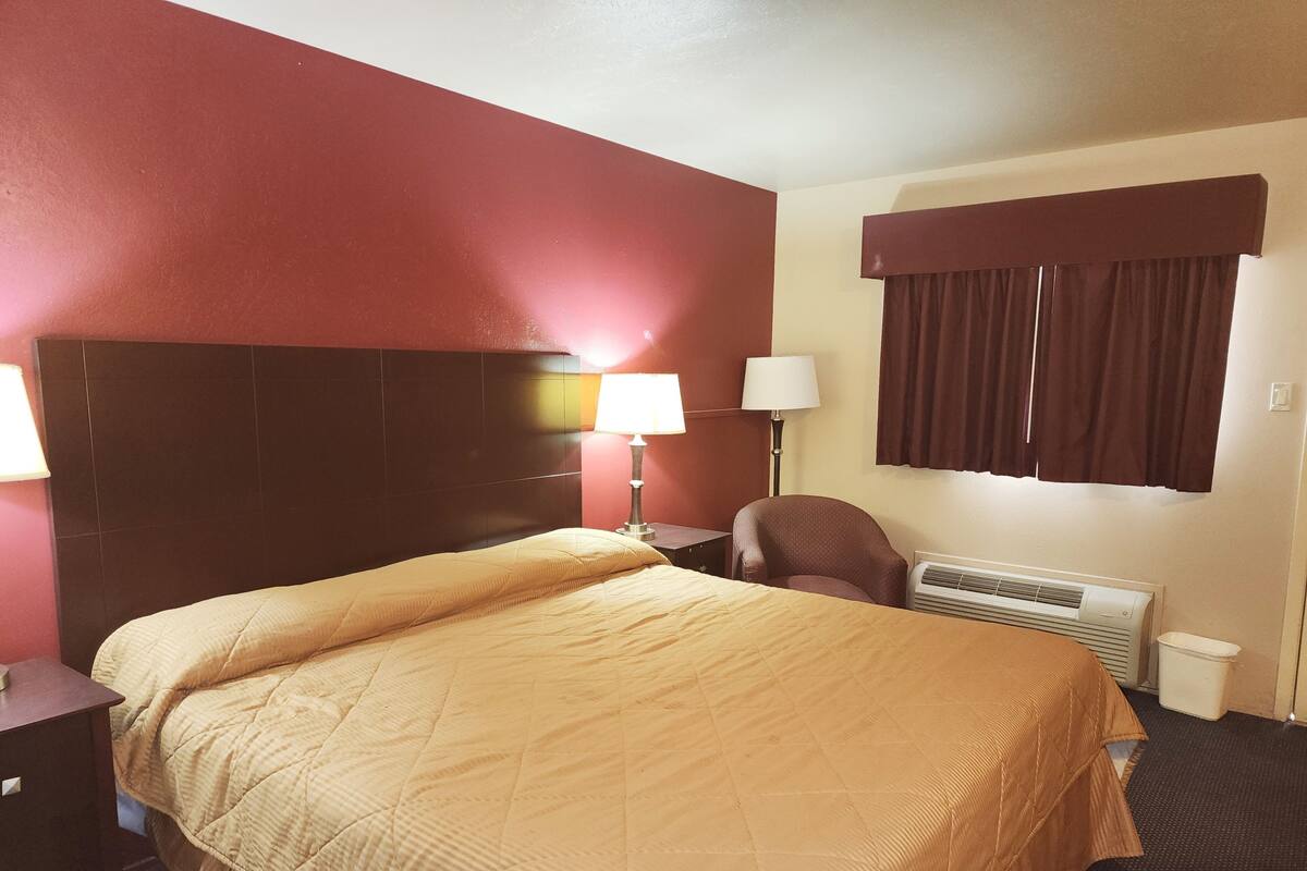 A hotel room with a bed, a chair, a lamp, a heater, and a window with curtains.