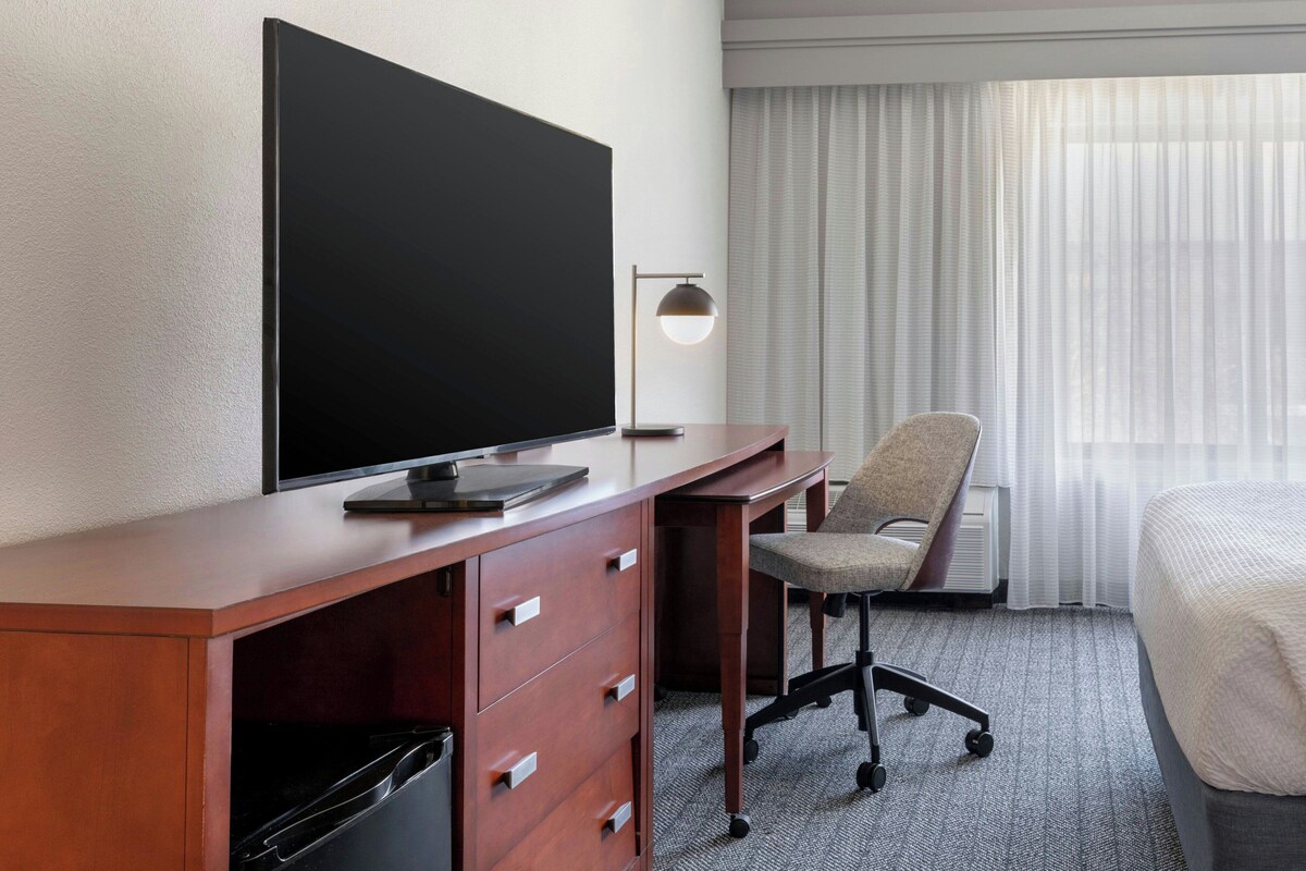 A hotel room with a desk, chair, TV, and a bed.