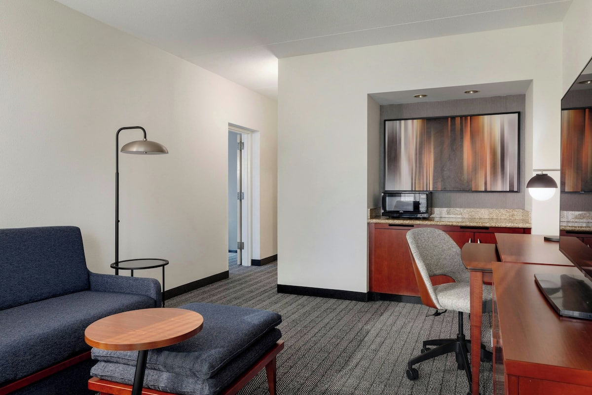 A hotel room with a sofa, a small round table, a desk with a chair, a kitchenette, and a large window.