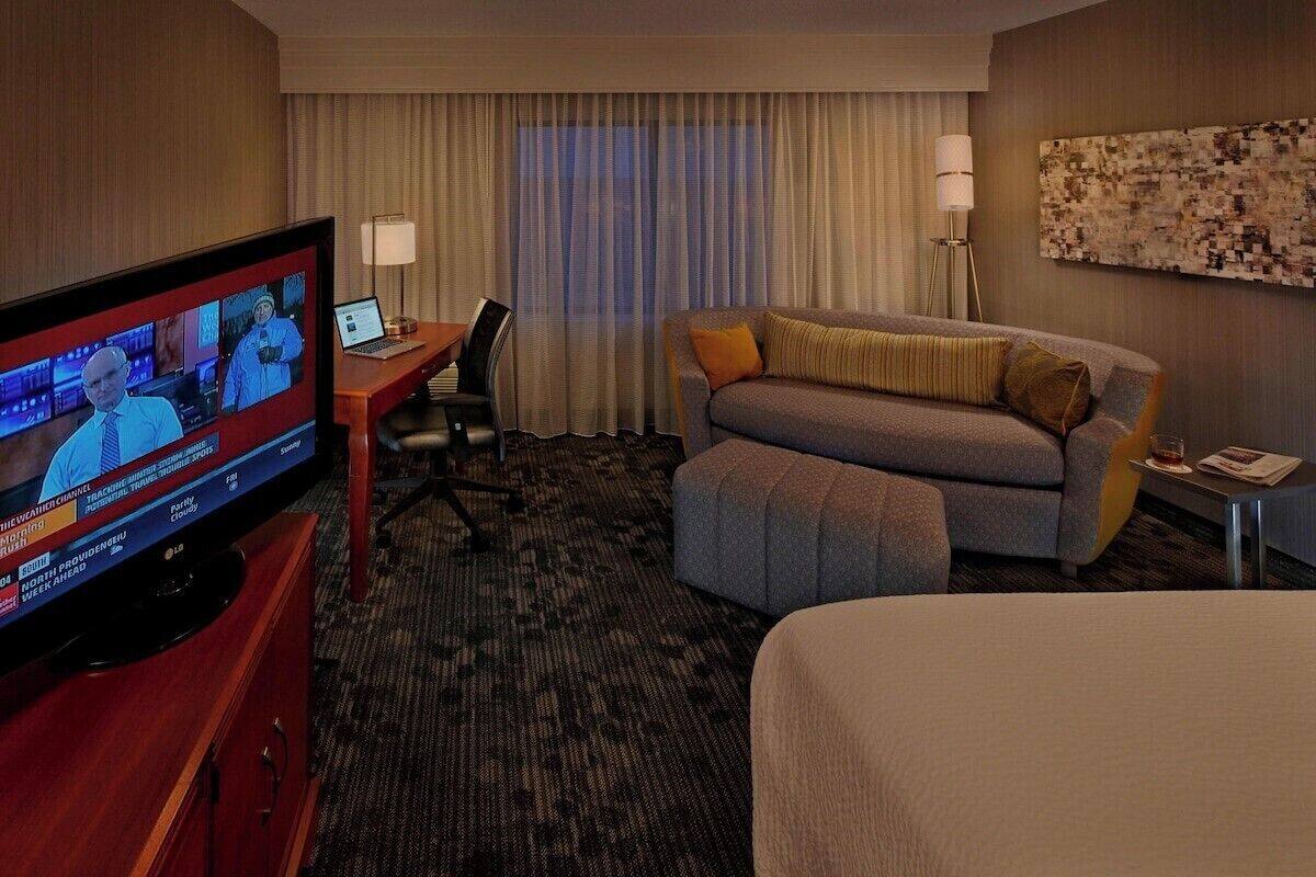 A hotel room with a television displaying news, a sofa, a desk with a laptop, and a lamp.
