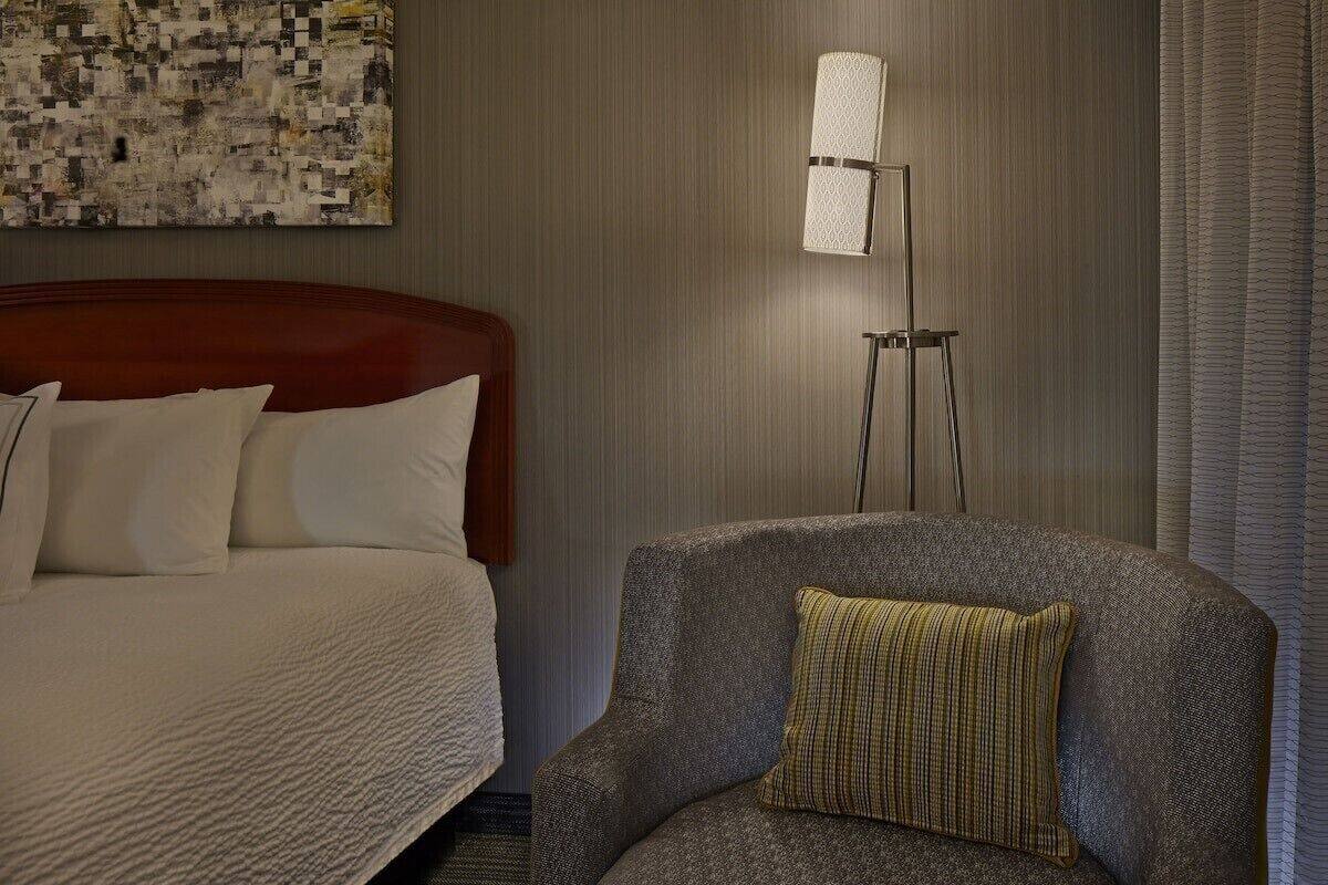 A hotel room with a bed, a chair, a lamp, and a wall-mounted artwork.