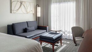 Una habitación de hotel con sofá, reposapiés, sillón y un cuadro en la pared.