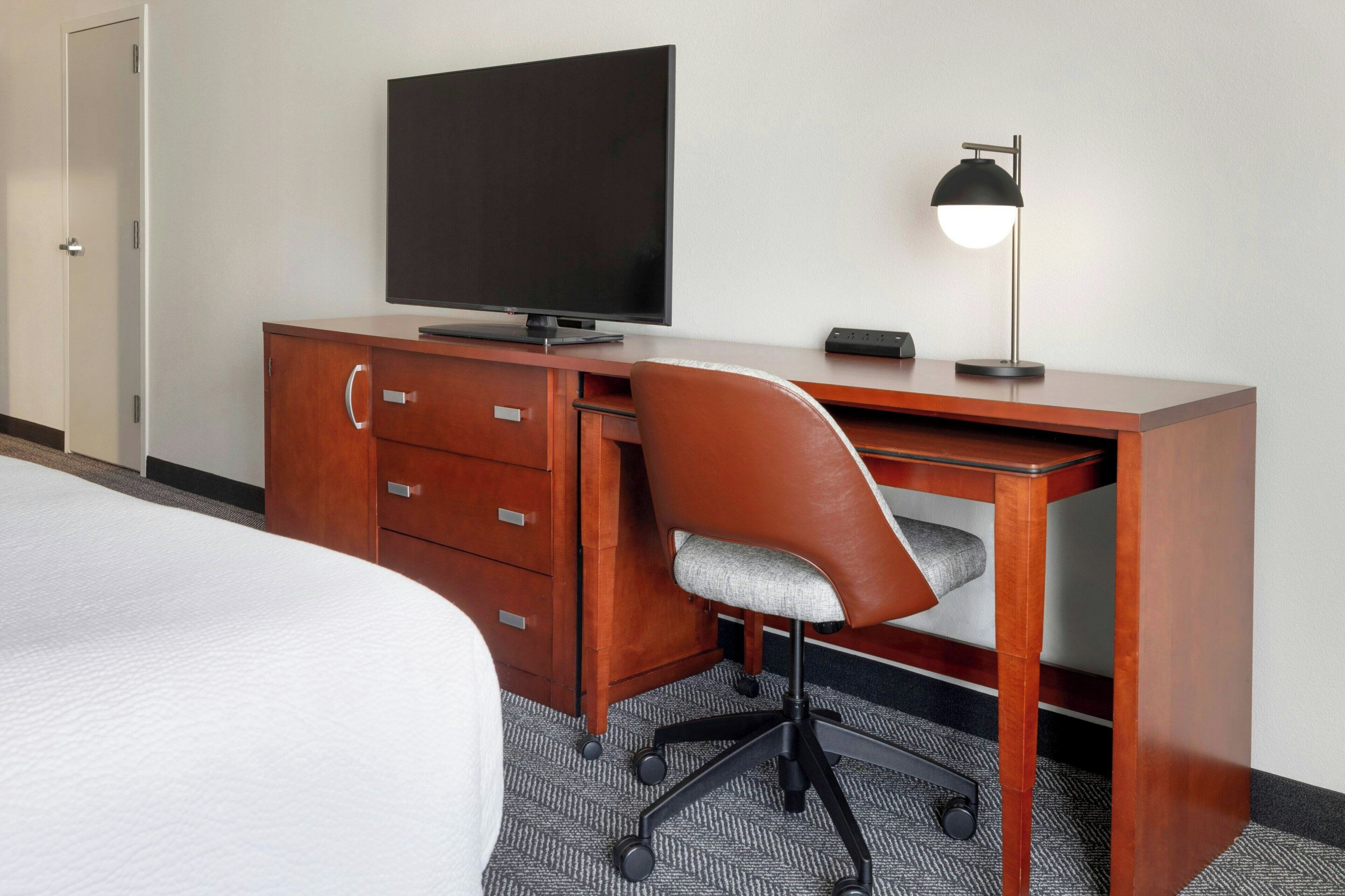 A hotel room with a bed, a wooden desk with a television, a chair, and a desk lamp.
