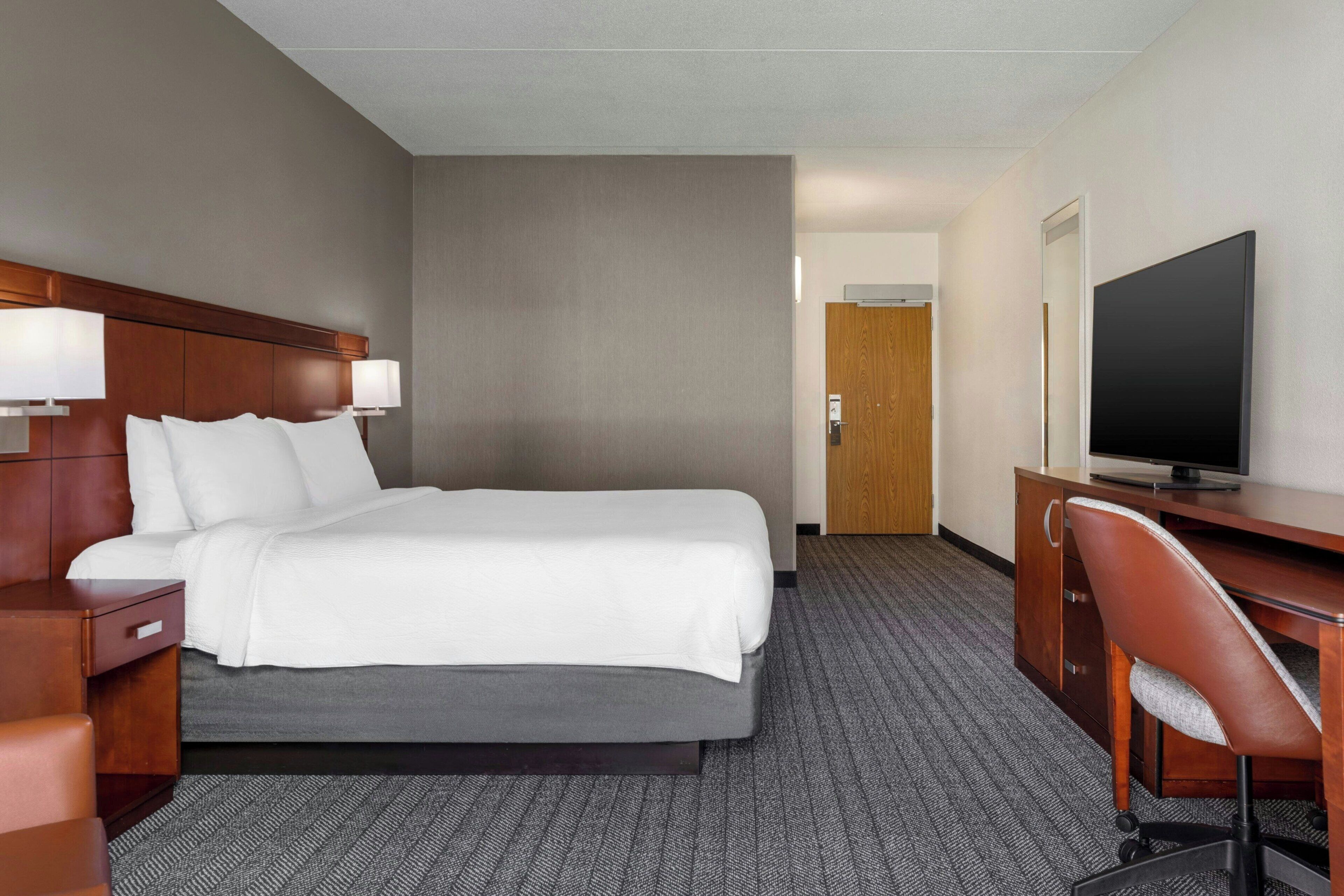 A hotel room with a bed, a desk with a chair, a television, and a door in the background.