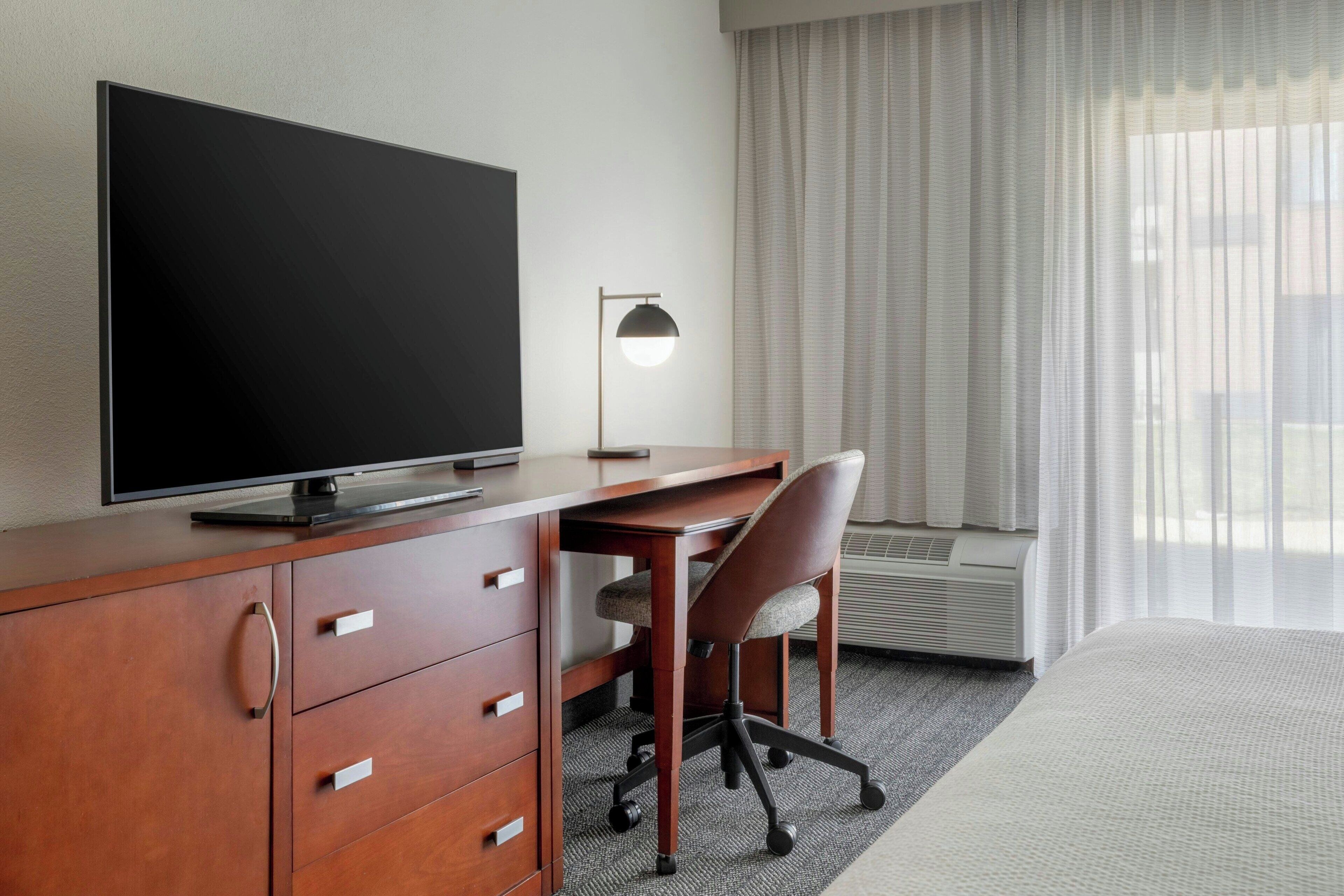 A hotel room with a bed, a desk with a chair, a television, a lamp, and a window with curtains.