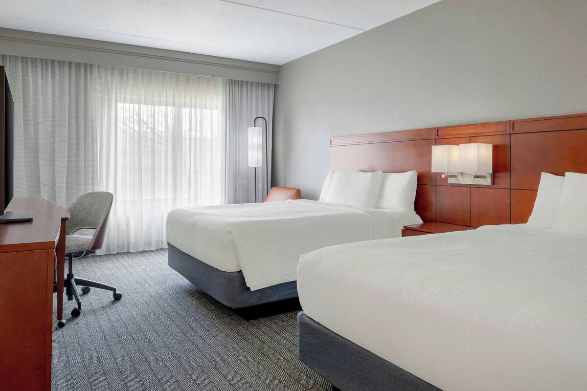 A hotel room with two beds, a desk, a chair, and a large window with curtains.
