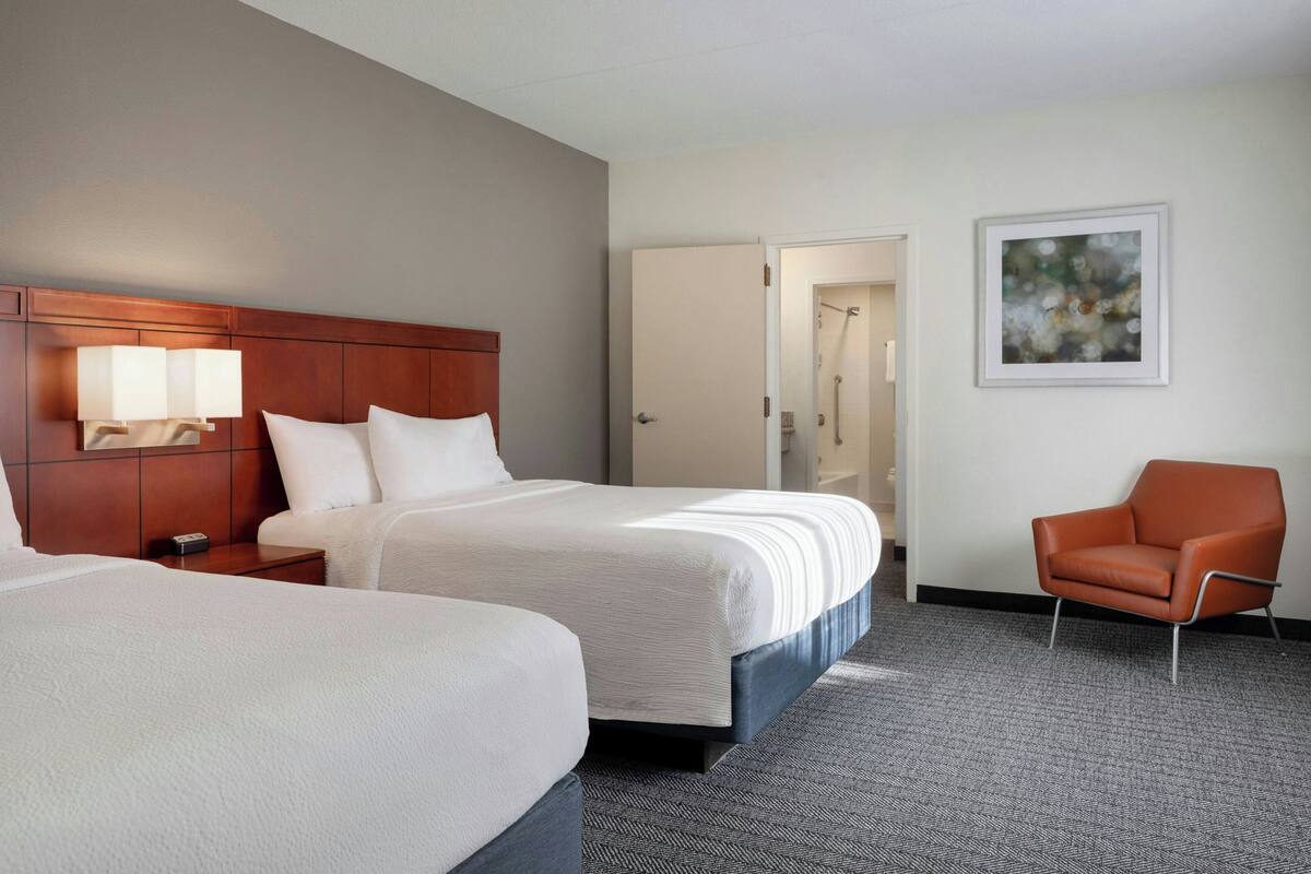 A hotel room with two beds, a chair, a painting, and a door leading to a bathroom.