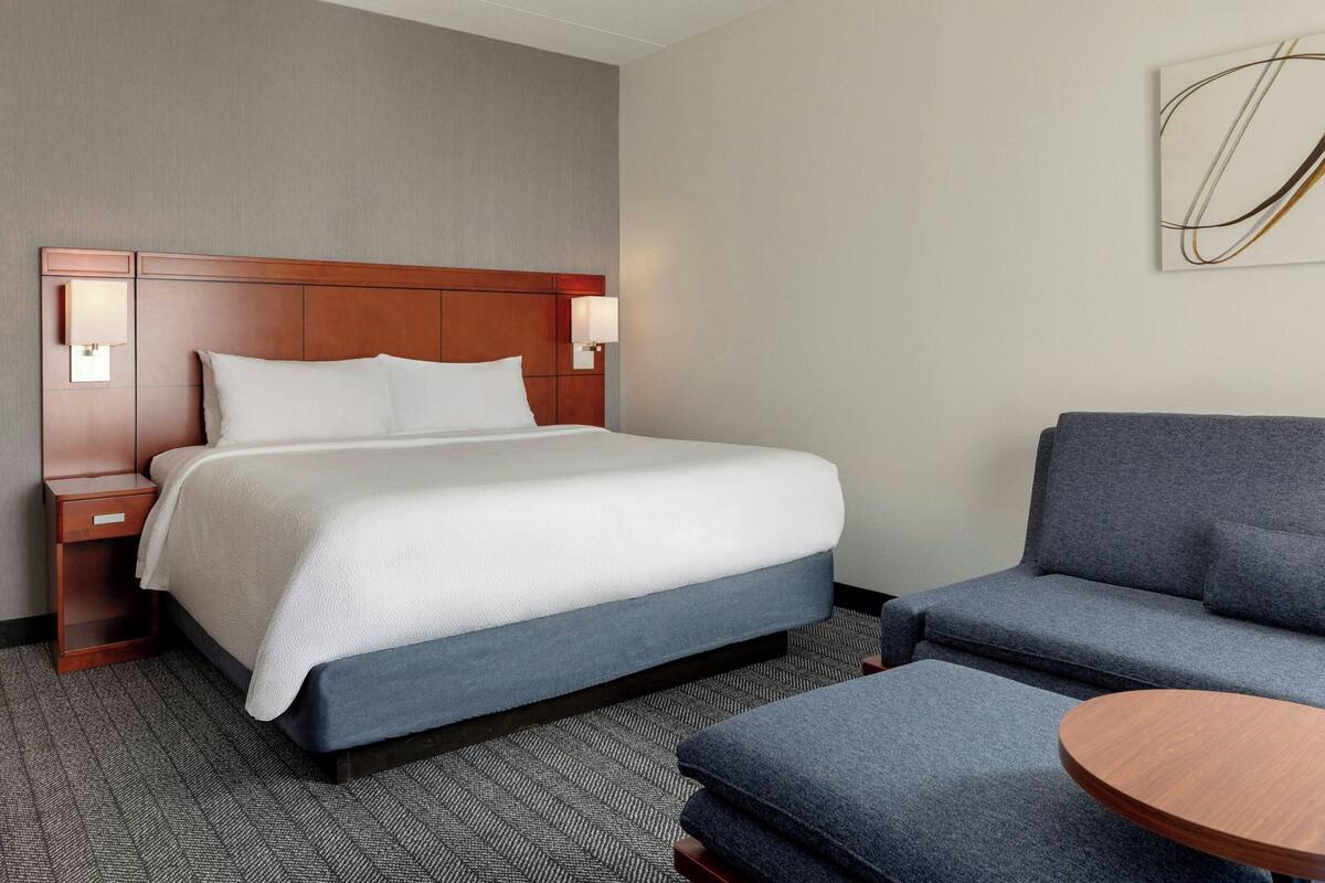 A hotel room with a bed, a sofa, a small table, and a wall-mounted lamp.