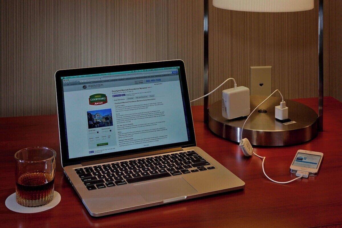A laptop on a wooden table with a glass of whiskey, a power strip, and a charging cable.
