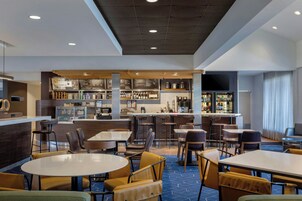 Un moderno vestíbulo de hotel con zona de bar, diferentes disposiciones de asientos y un cartel de menú visible.