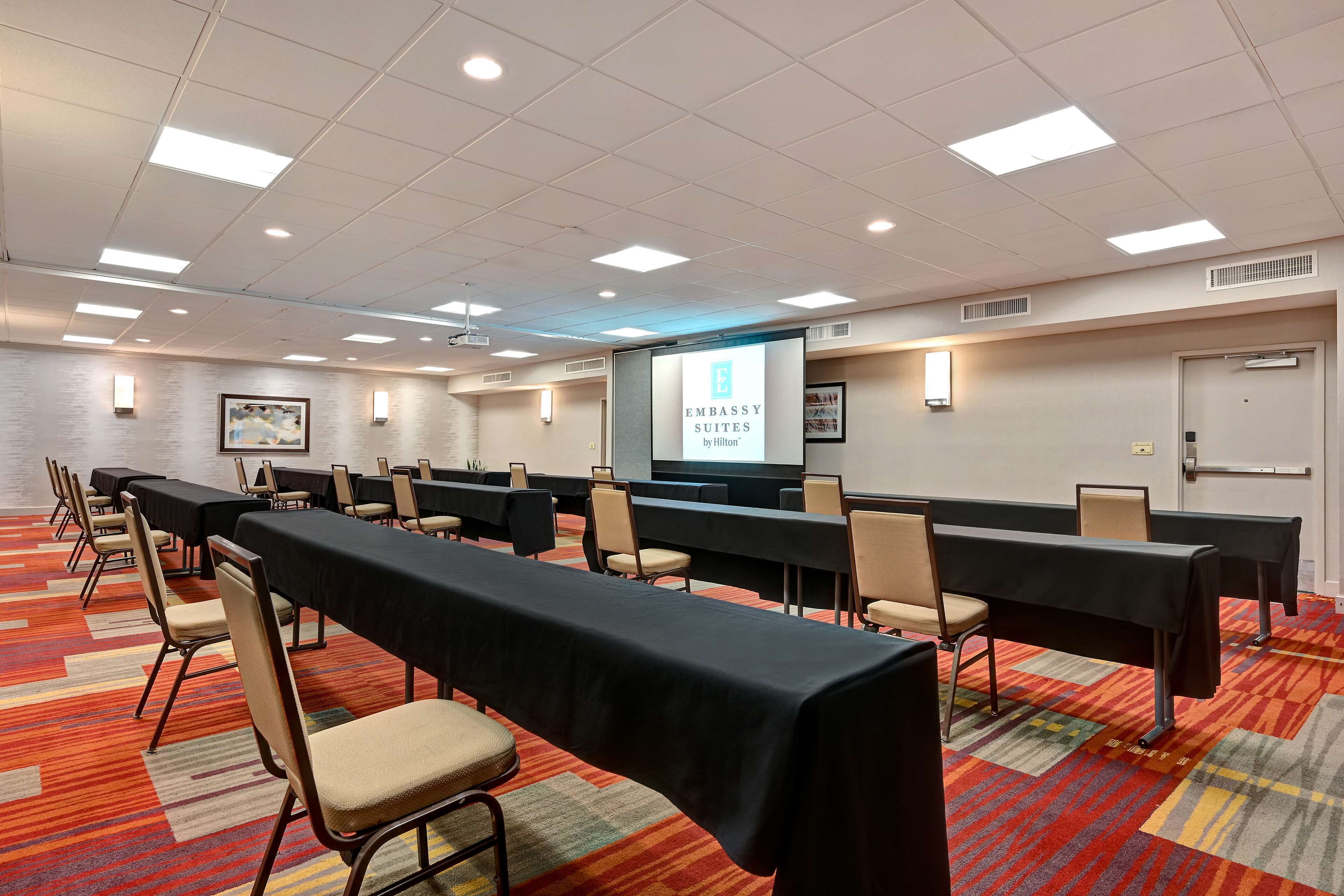 A conference room with long tables, chairs, a projector screen, and a carpeted floor.