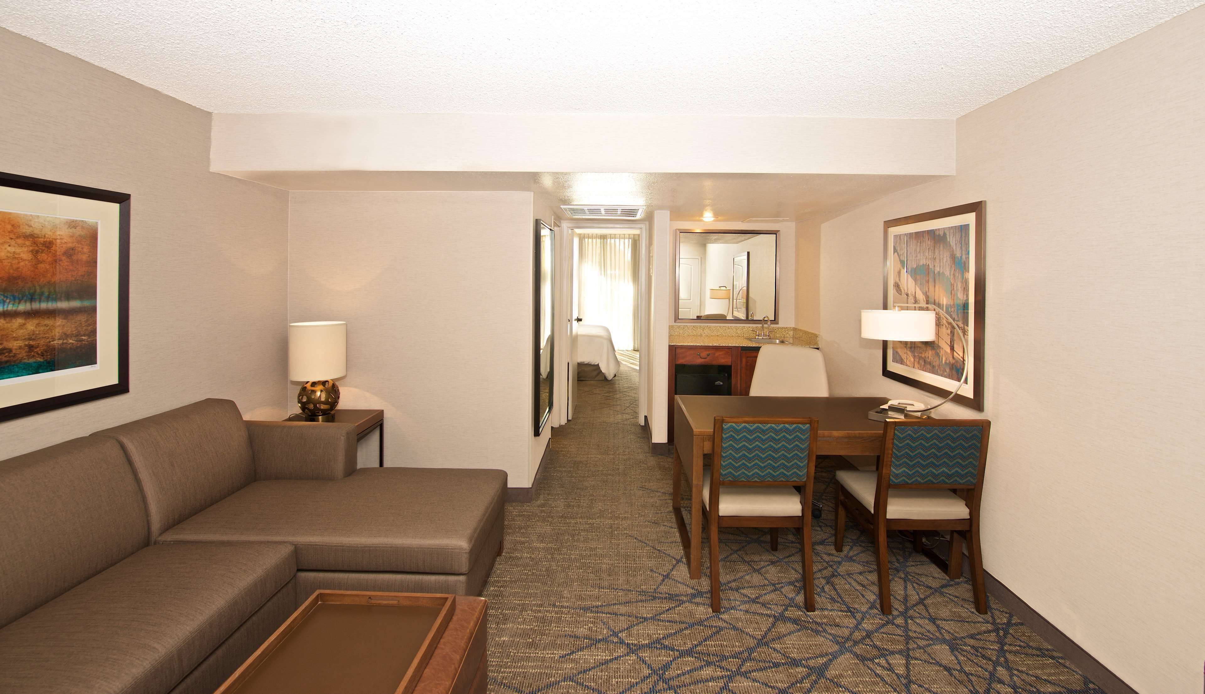 A hotel room with a sofa, a dining table with chairs, a desk, a mirror, and a painting on the wall.
