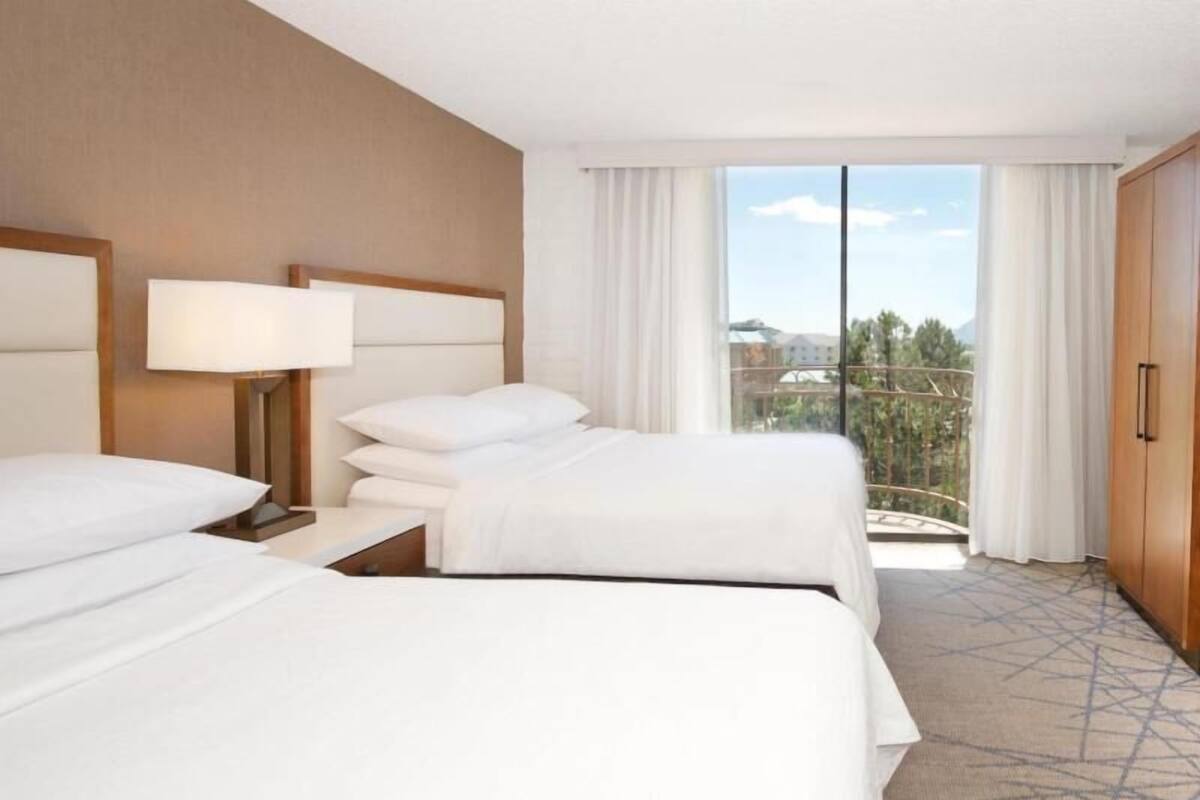 A hotel room with two beds, a nightstand with a lamp, a wardrobe, and a large window with a view of buildings and trees.