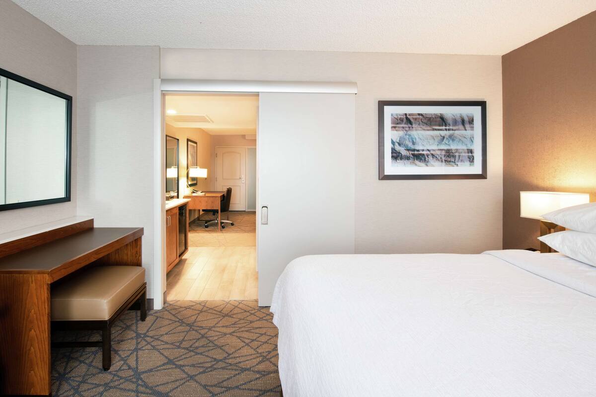 A hotel room with a bed, a desk with a chair, a bathroom with a mirror, and a framed picture on the wall.