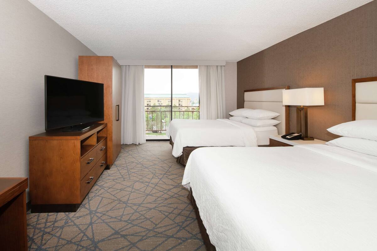 A hotel room with a television on a wooden cabinet, a bed with white bedding, a bedside table with a lamp, and a window with curtains.