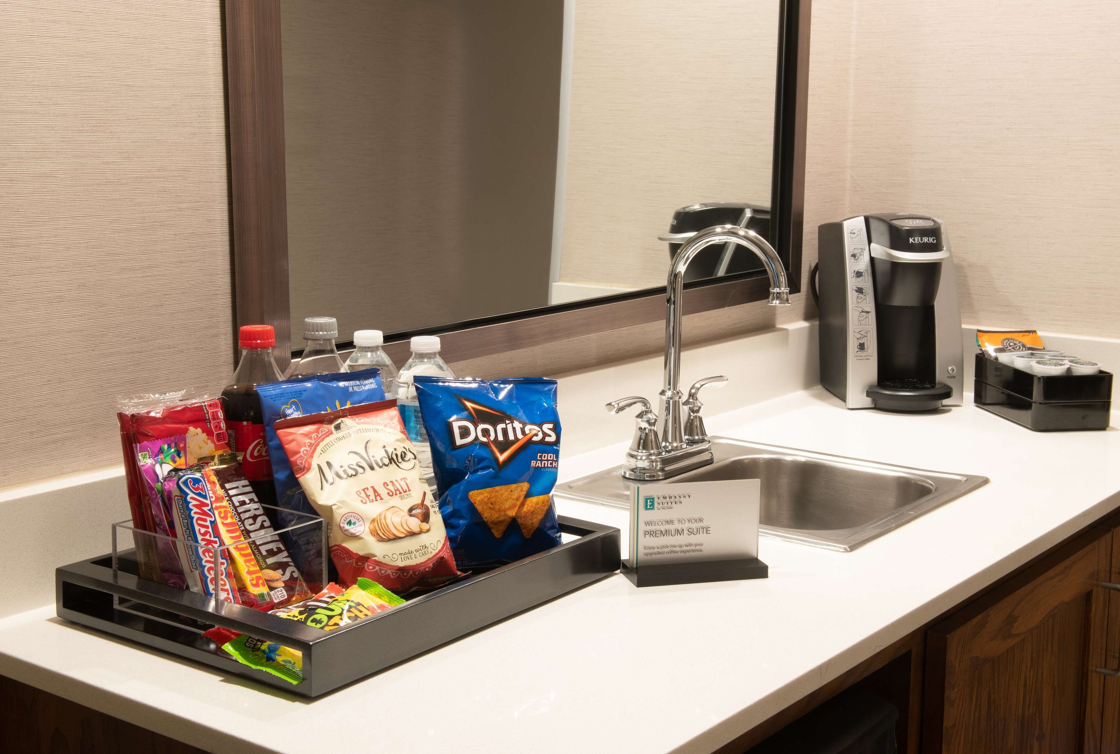A hotel bathroom counter with a coffee maker, a sink, and snack dispensers including chips and candy.