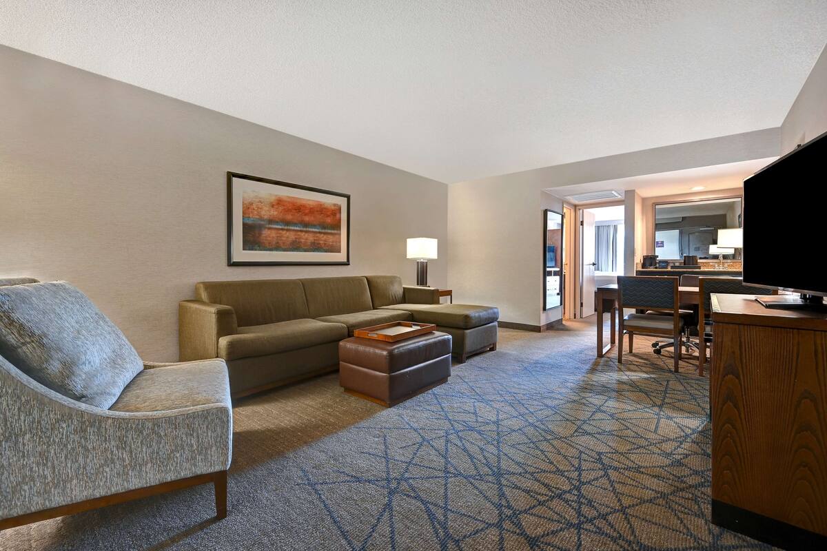 A hotel room with a sofa, armchair, ottoman, and a desk with a television.
