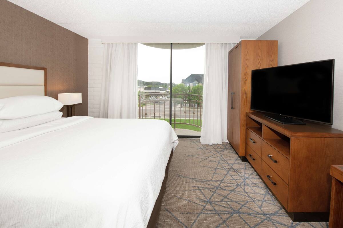 A hotel room with a large bed, a TV on a wooden cabinet, and a balcony with a view of the city.