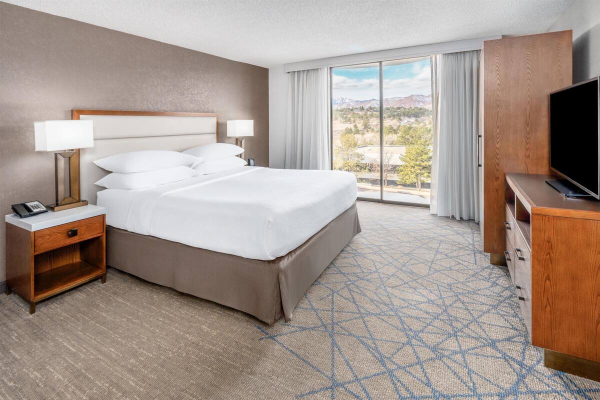 A hotel room with a large bed, two bedside tables with lamps, a TV, and a view of mountains through the window.