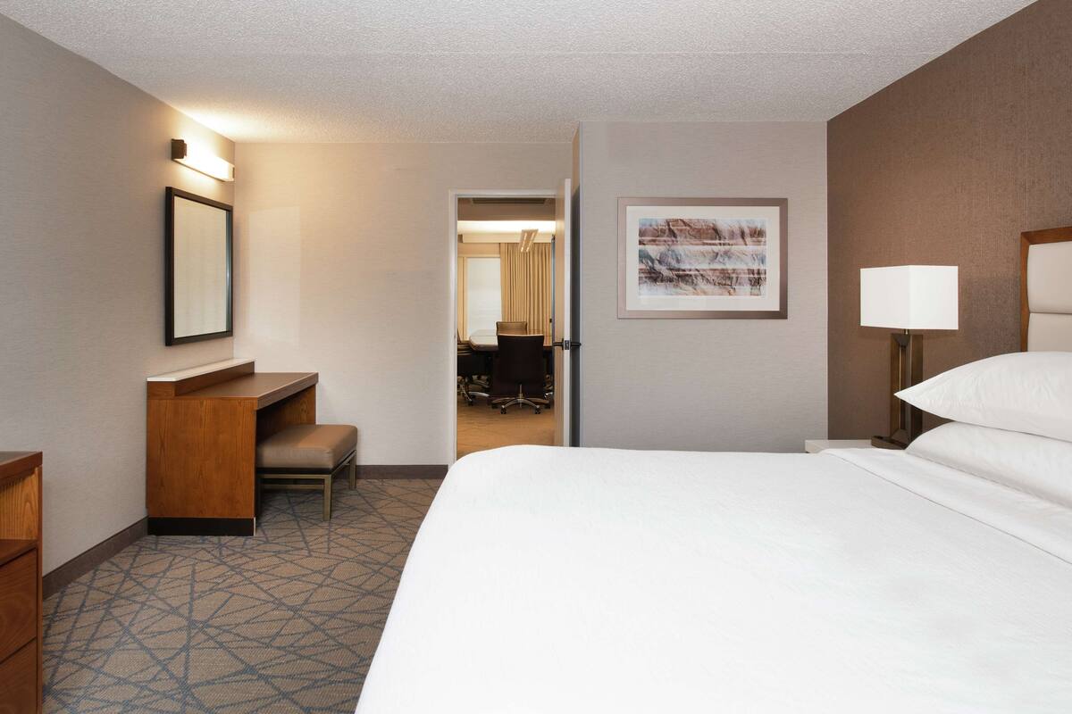 A hotel room with a large bed, a desk with a chair, a small table, a lamp, and a framed picture on the wall.