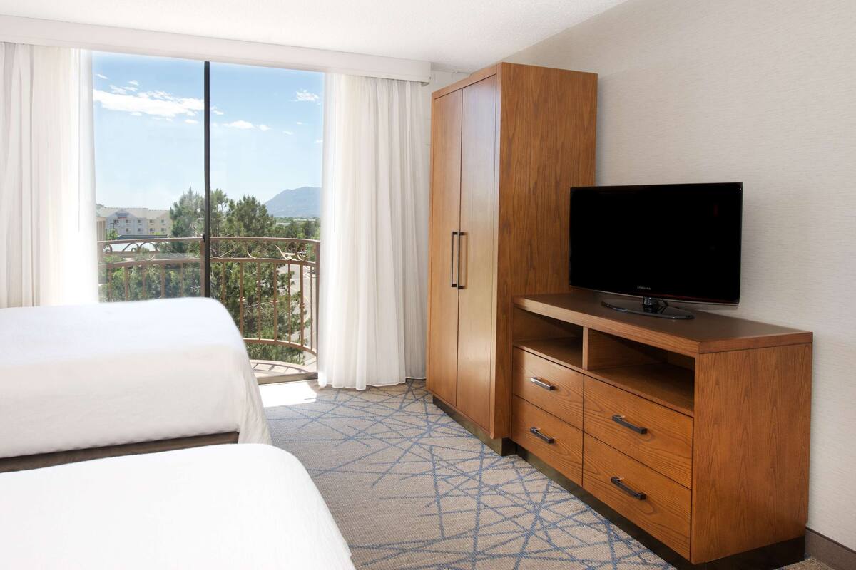 A hotel room with a bed, a TV on a wooden stand, and a balcony with a view of the city and mountains.